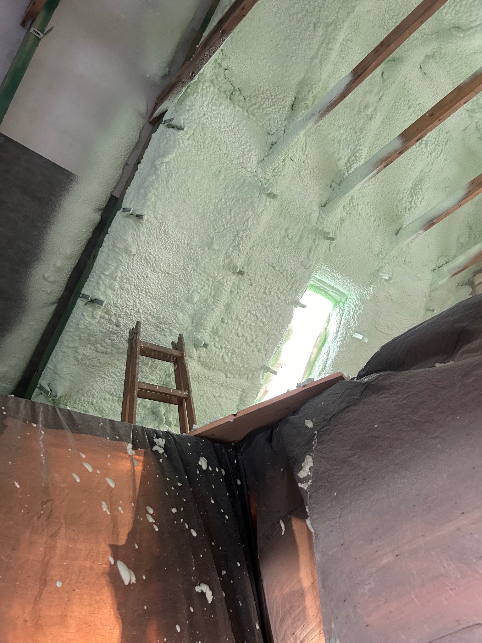 Poddasze z drewnianą konstrukcją pokryte warstwą jasnej pianki izolacyjnej, widoczne okno dachowe i drabina oparta o ścianę, folia ochronna na elementach konstrukcyjnych.