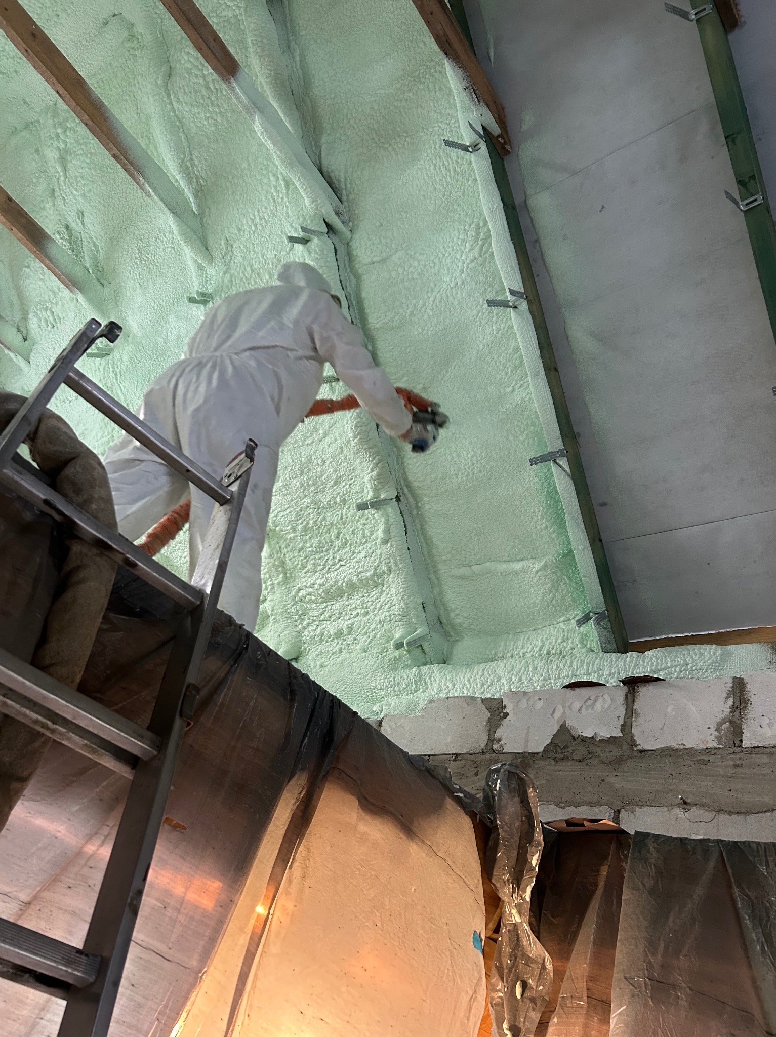 Pracownik w kombinezonie ochronnym natryskuje pianę izolacyjną na poddasze z drewnianą konstrukcją, widoczny fragment drabiny i folii ochronnej.