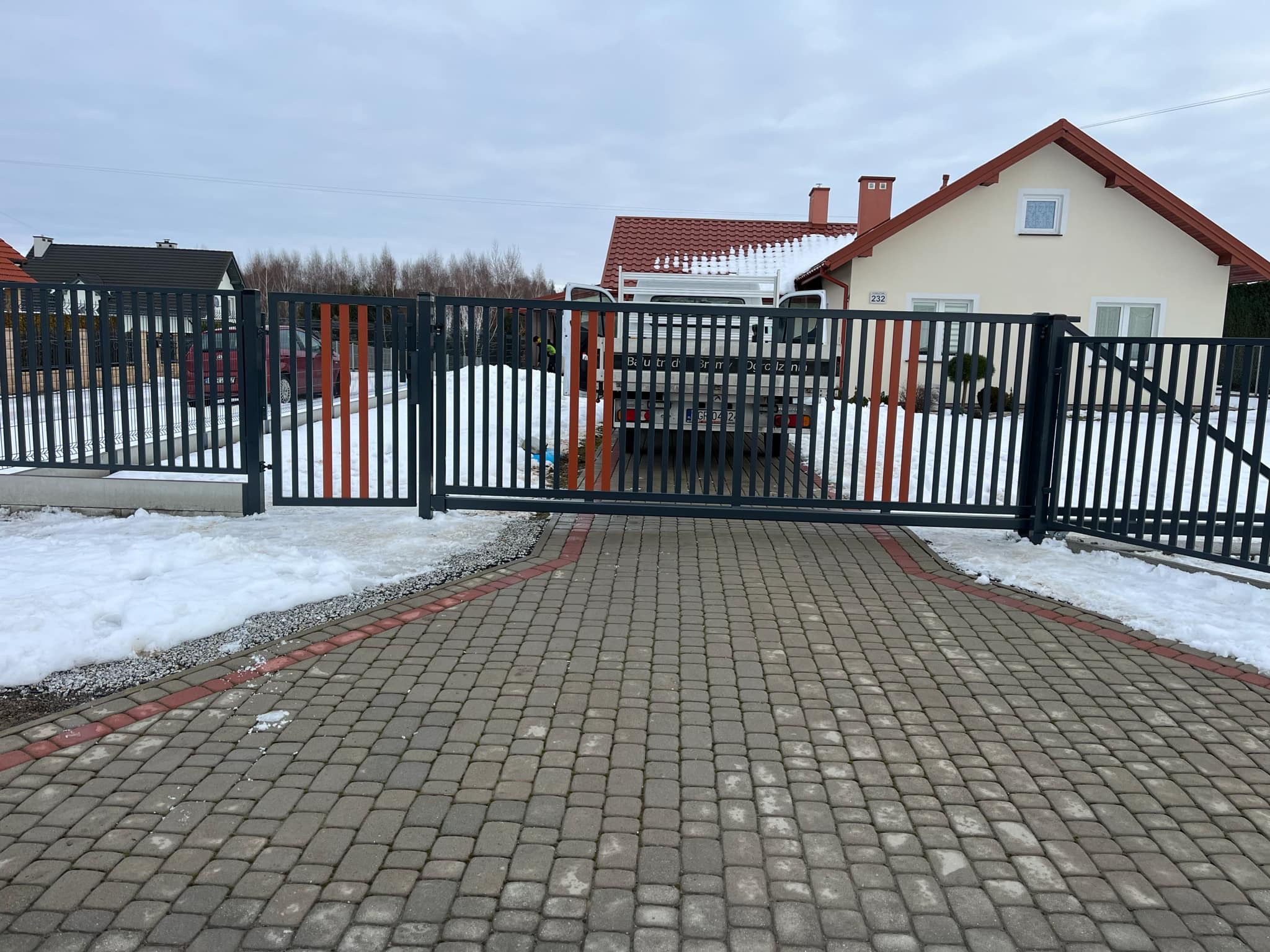 Nowoczesna, przesuwna brama wjazdowa z metalowymi sztachetami w kolorze grafitowym, z pomarańczowymi akcentami, na tle domu z czerwonym dachem i podjazdu z kostki brukowej, w zimowej scenerii.