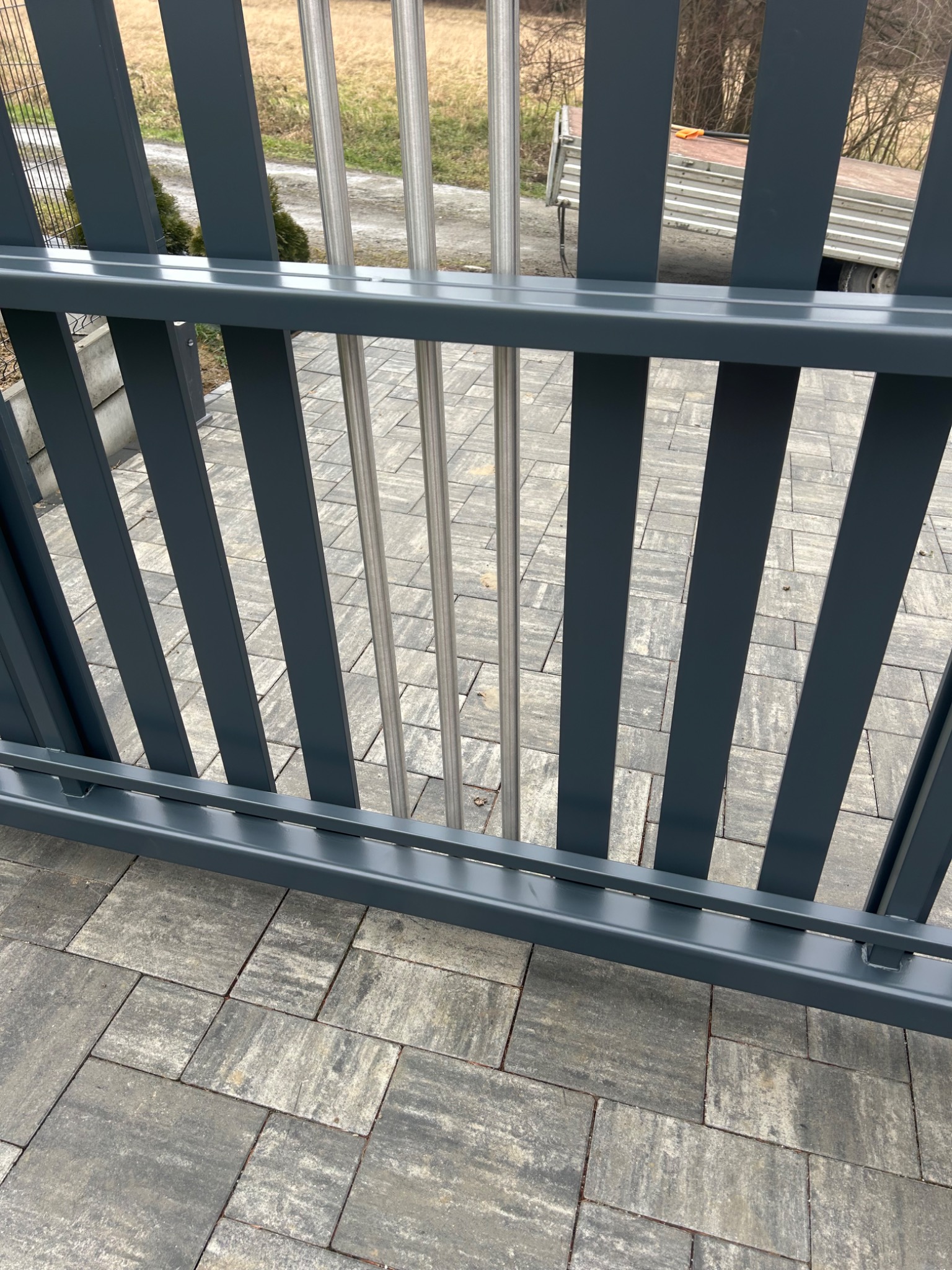 Nowoczesna brama wjazdowa z szarymi, pionowymi panelami i trzema srebrnymi prętami, widoczna z bliska na tle kostki brukowej i przyczepy.