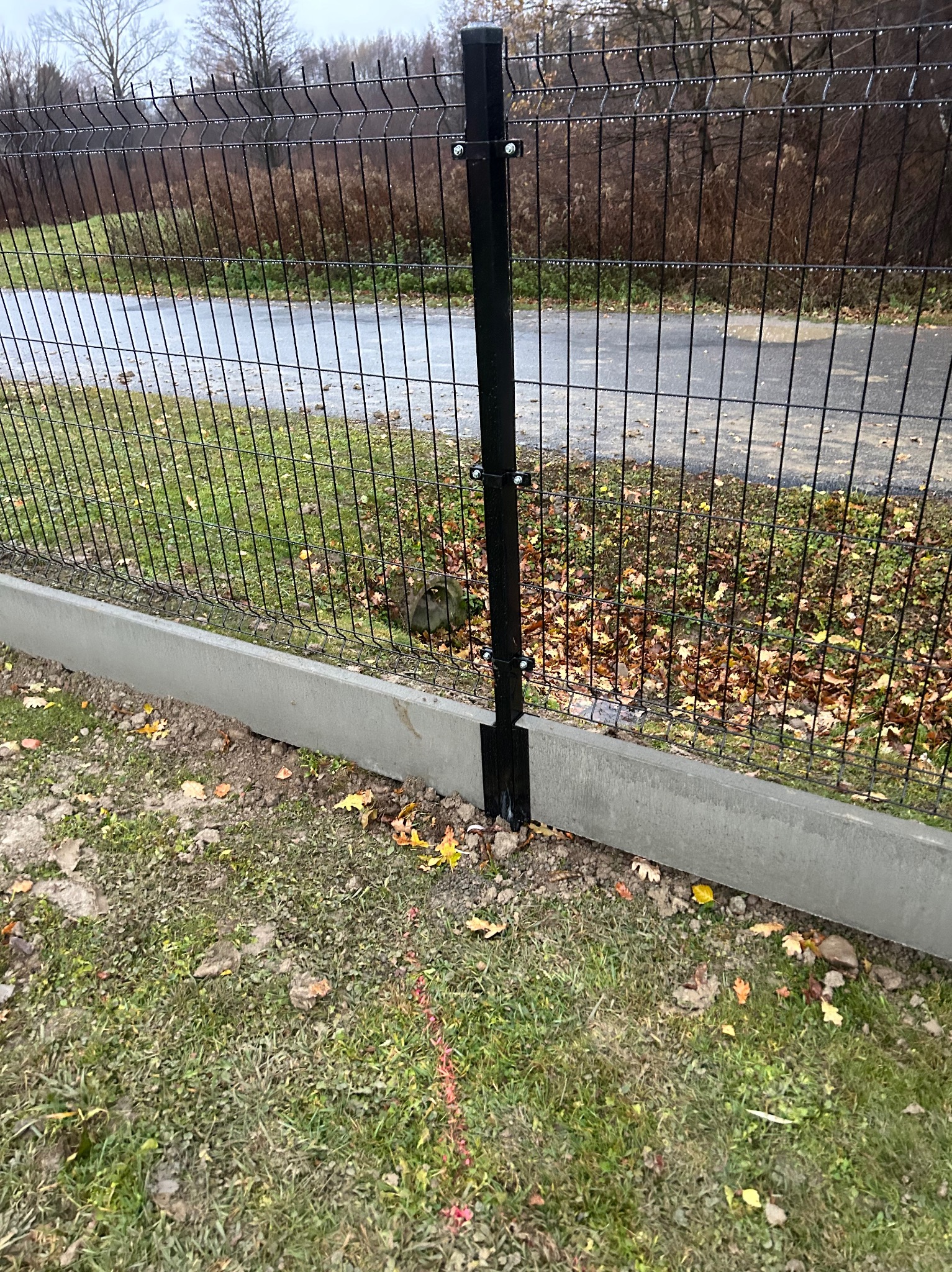 Fragment czarnego ogrodzenia panelowego z podmurówką betonową, widok z bliska na słupek i przęsło, z widoczną linią wytyczającą przebieg ogrodzenia na trawie.