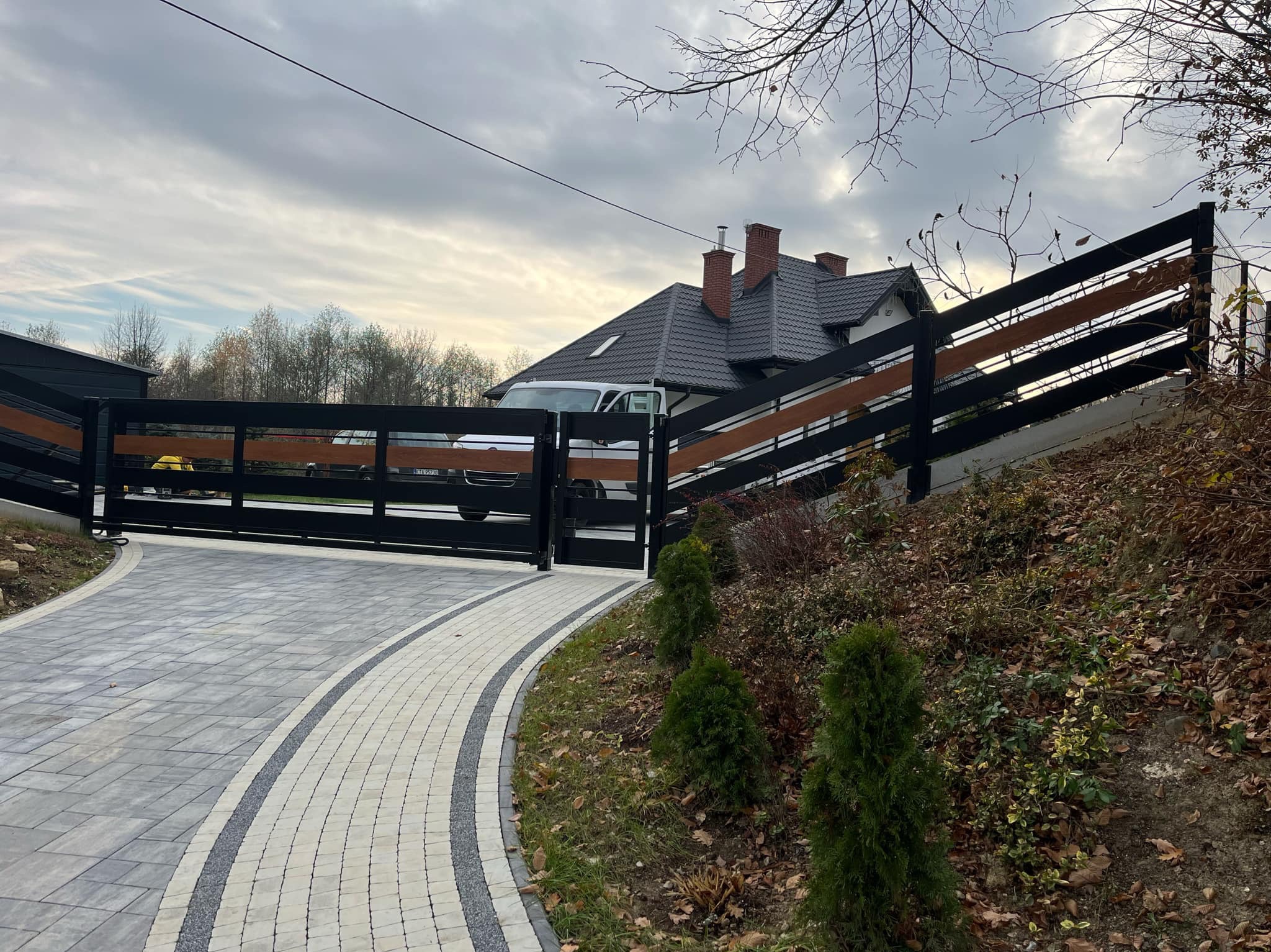 Nowoczesna, przesuwna brama wjazdowa z czarnymi metalowymi elementami i poziomymi panelami z drewna, widoczna z zewnątrz posesji na tle domu z ciemną dachówką i podjazdem z kostki brukowej.