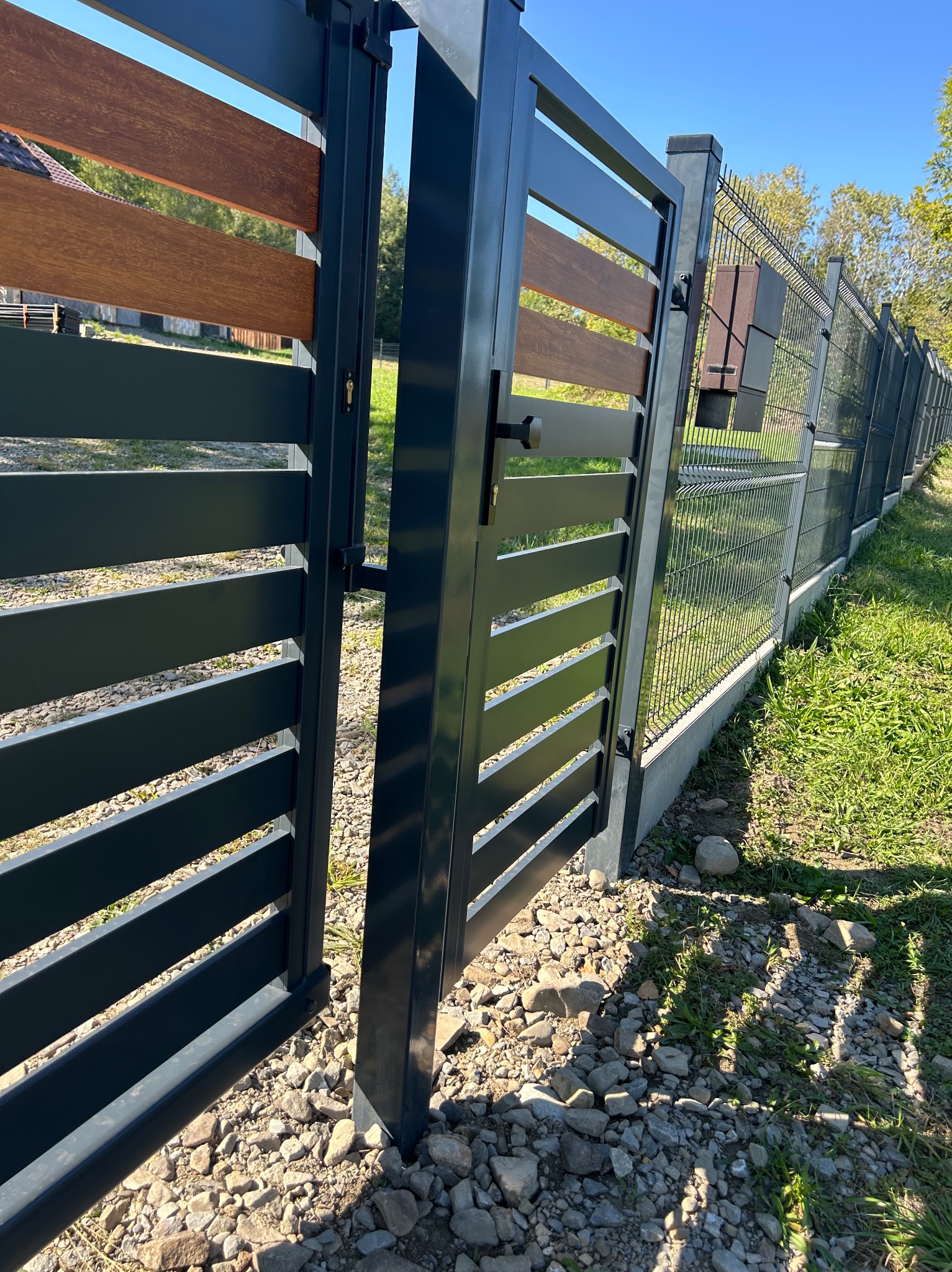 Otwarta, nowoczesna brama wjazdowa z poziomymi, drewnopodobnymi panelami w antracytowej ramie, sąsiadująca z ogrodzeniem z siatki na betonowej podmurówce, widok z poziomu gruntu.