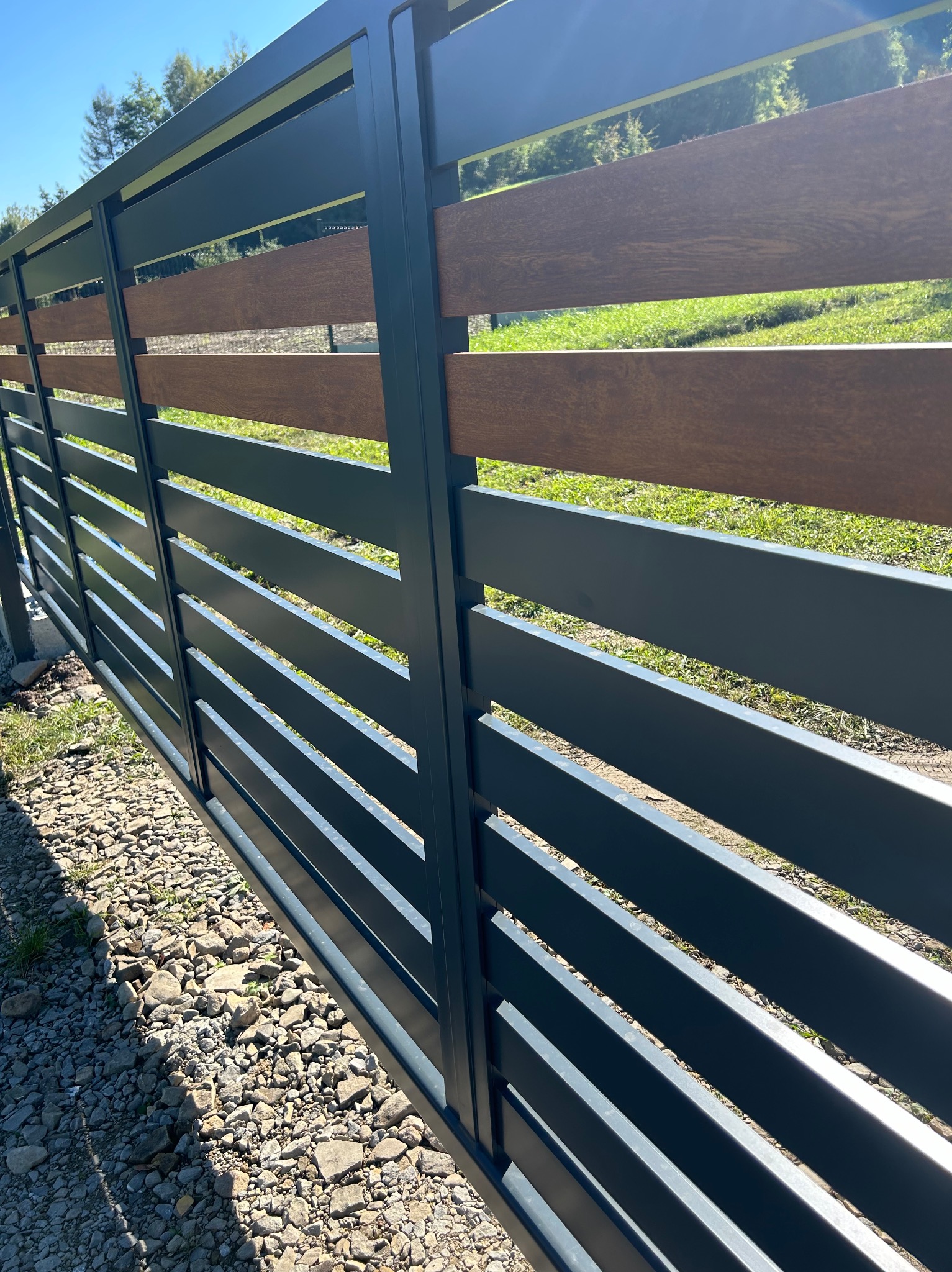 Nowoczesne ogrodzenie z poziomych, szarych paneli metalowych i wstawkami z drewna kompozytowego, widok z zewnątrz na tle zielonego trawnika i drzew, żwirowe podłoże u podstawy.