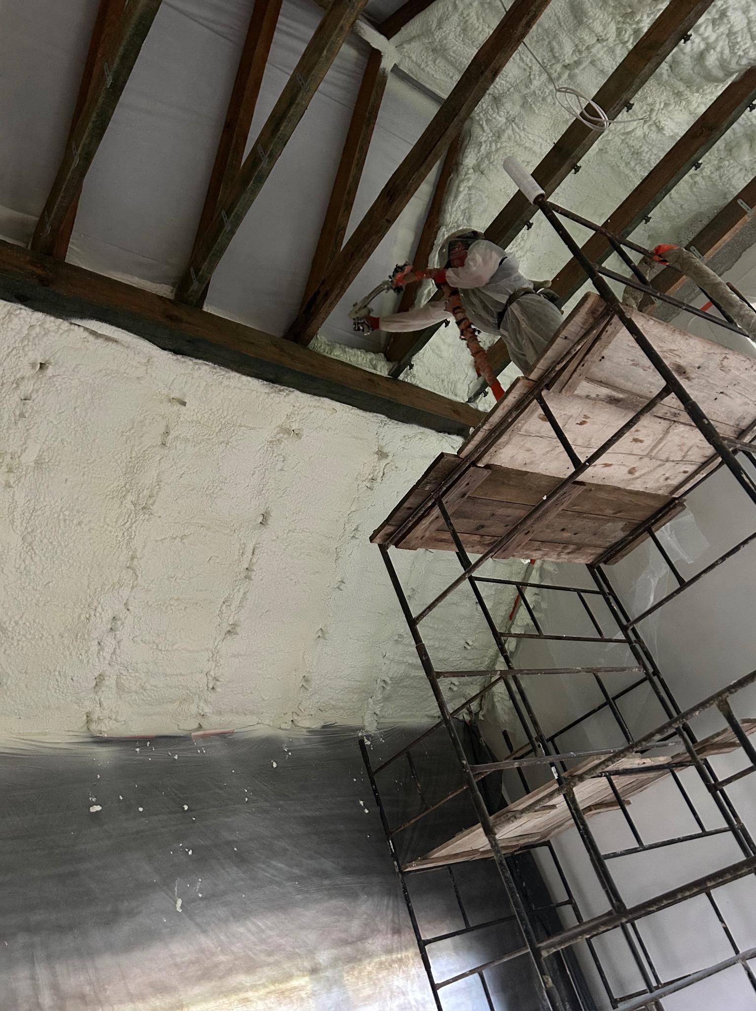 Praca przy ocieplaniu poddasza pianką PUR w Uściu Gorlickim. Widok z dołu na rusztowanie, pracownika w kombinezonie i nałożoną pianę na konstrukcji dachu.