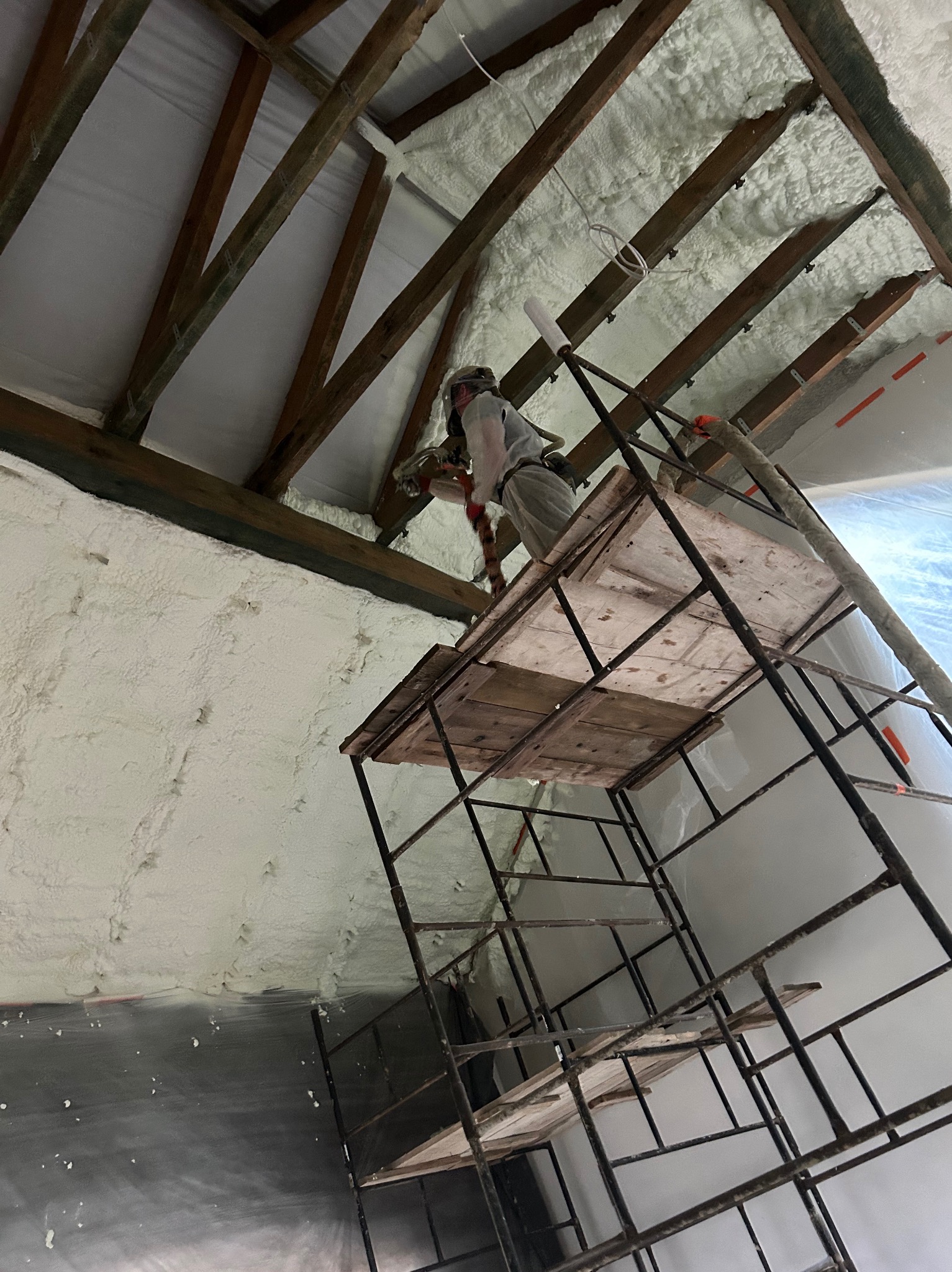 Pracownik w kombinezonie ociepla poddasze pianką PUR, stojąc na rusztowaniu. Widoczne drewniane elementy konstrukcyjne dachu i zaizolowana powierzchnia.