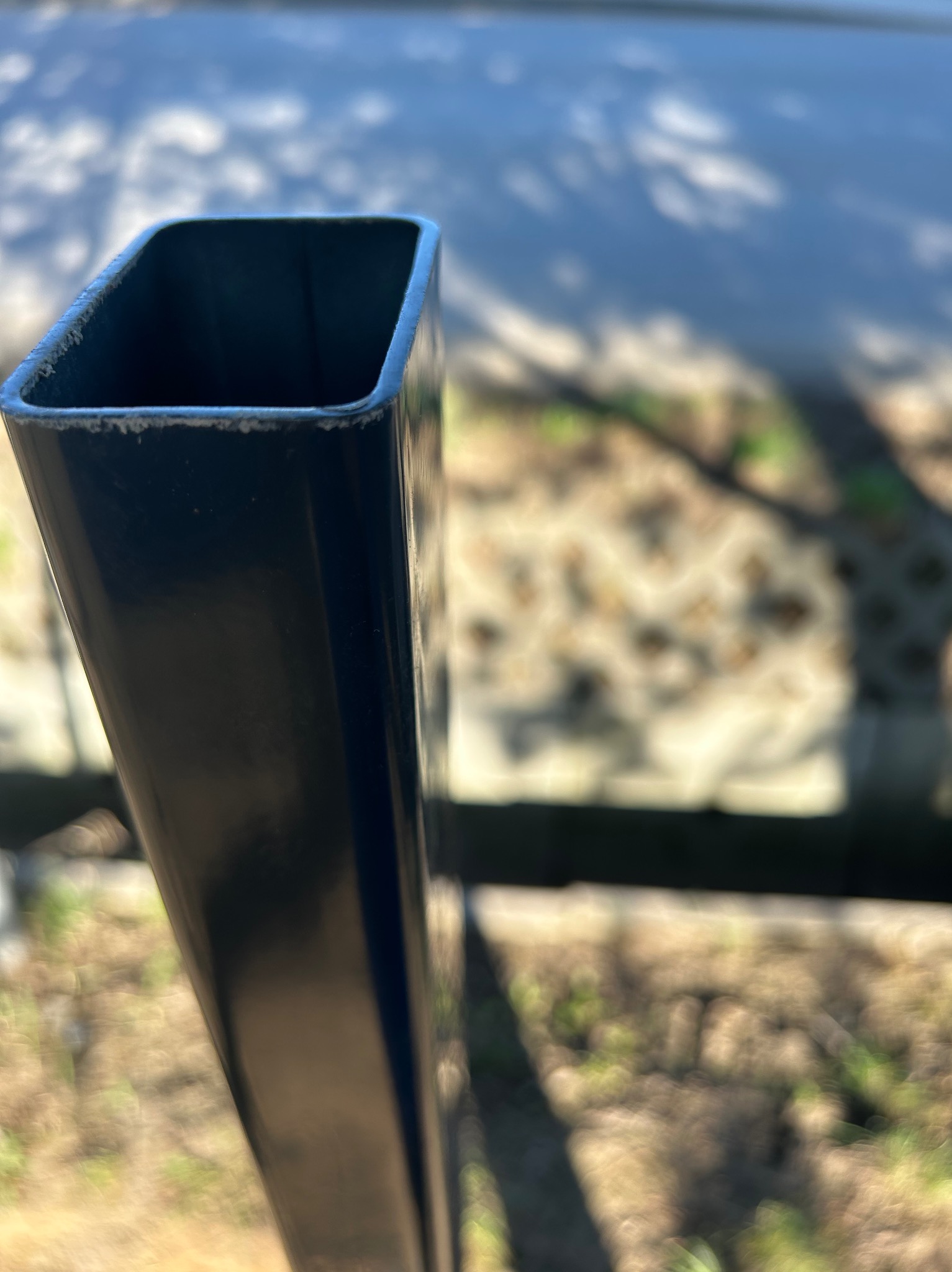 Metalowy słupek ogrodzeniowy o kwadratowym przekroju, widoczny z bliska na tle rozmytego otoczenia. Słupek jest czarny, a tło stanowią elementy ogrodzenia i zieleń.