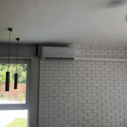 Klima-Gold Łukasz Olejnik - Biały klimatyzator AUX zamontowany na ścianie z białej cegły w jasnym wnętrzu. Widoczne okno i lampy wiszące.