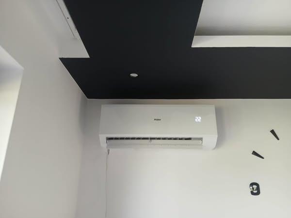 Biała klimatyzacja Haier na ścianie z dekoracyjnym zegarem. Czarny, geometryczny wzór na suficie. Minimalistyczny design wnętrza, nowoczesne wykończenie.