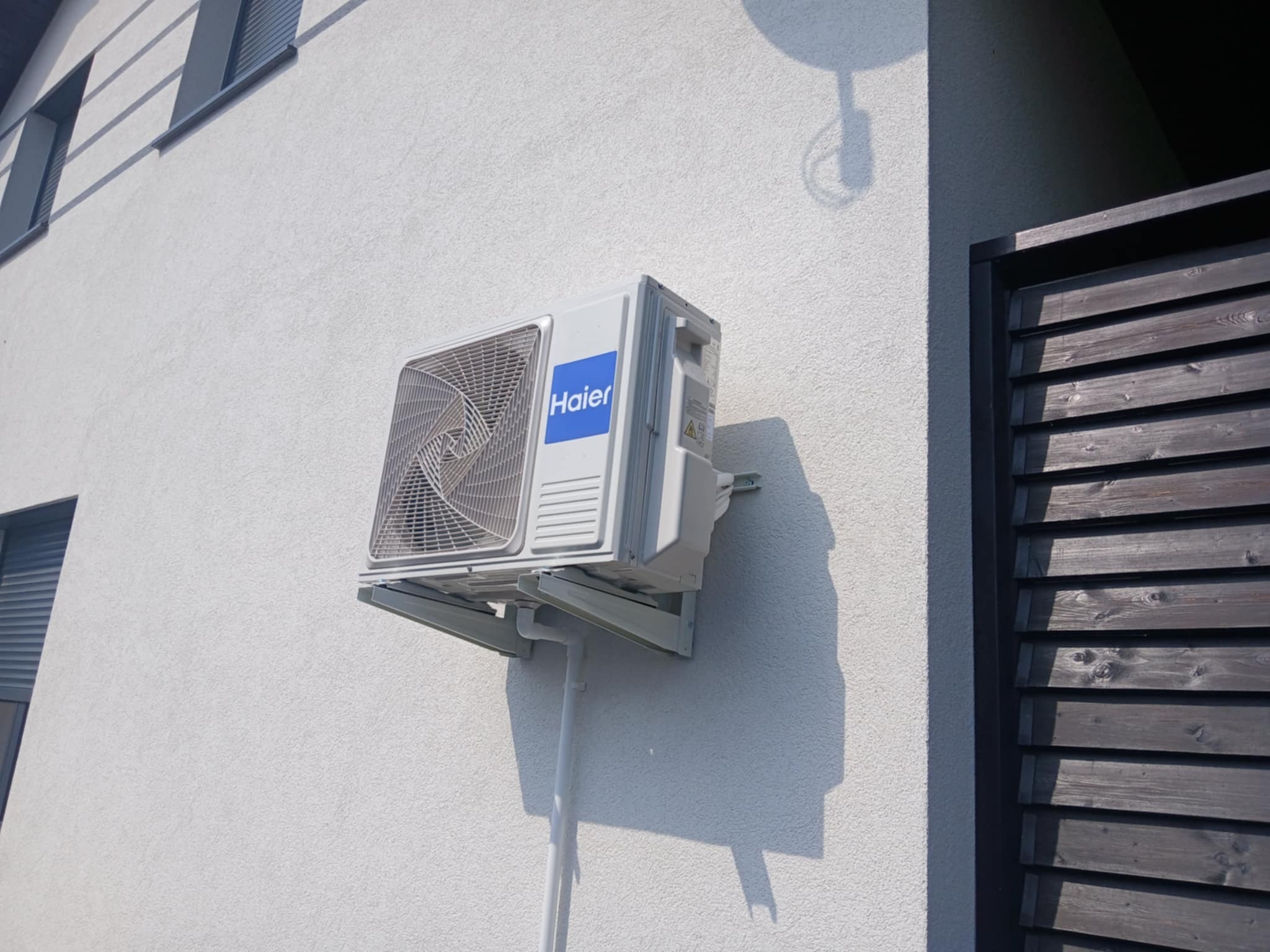 Biały klimatyzator Haier zamontowany na jasnej, teksturowanej ścianie budynku, z widocznymi elementami elewacji i instalacji odprowadzającej skropliny.