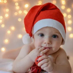 Uroczy niemowlak w czerwonym czepku Mikołaja, leżący na białym tle z lampkami, trzymający czerwoną bombkę. Wyjątkowe ujęcie świąteczne, idealne na pamiątkę.