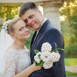 Studiyasveta - Uśmiechnięta para młoda w objęciach, panna młoda w koronkowej sukni i welonie, pan młody w ciemnym garniturze trzyma bukiet białych róż na tle zieleni.