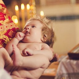 Studiyasveta - Niemowlę w ramionach osoby w bogato zdobionej szacie liturgicznej, prawdopodobnie podczas ceremonii chrztu, w tle świece i ikona.