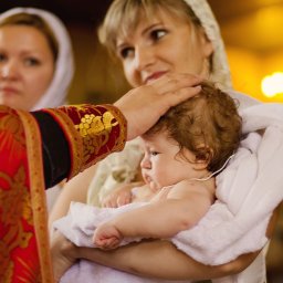 Studiyasveta - Chrzest dziecka w cerkwi: matka trzyma niemowlę owinięte w biały ręcznik, ksiądz w bogato zdobionej szacie błogosławi, w tle kobieta w chuście.