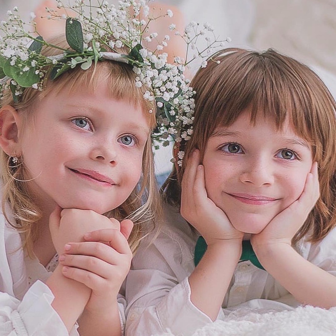 Uśmiechnięte dzieci: blondynka z wiankiem z gipsówki i brunet w zielonej muszce, opierają twarze na dłoniach, leżąc na białym tle. Portret w naturalnym świetle.