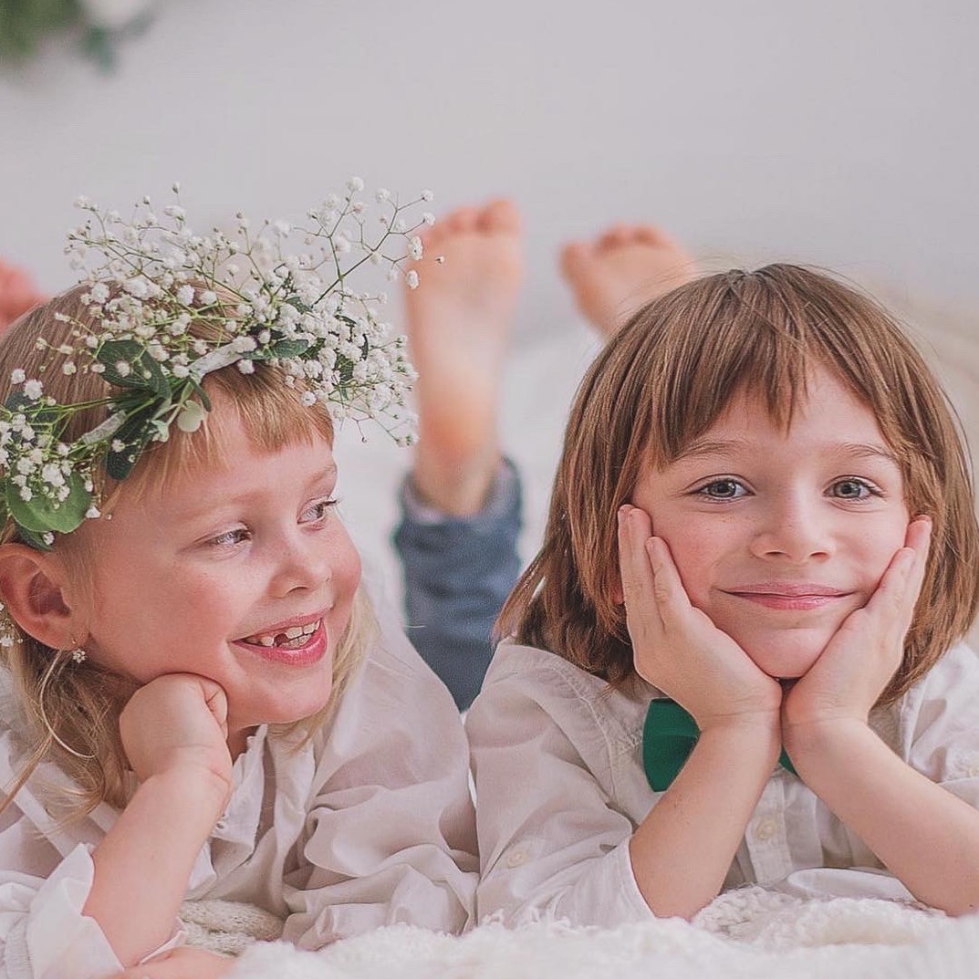 Uśmiechnięte rodzeństwo leżące na białym kocu: dziewczynka z wiankiem z gipsówki i chłopiec w zielonej muszce. Ujęcia z perspektywy bliskiej, delikatne oświetlenie.