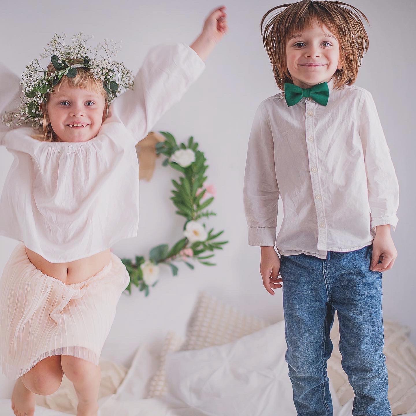 Dzieci w skoku na łóżku, dziewczynka z wiankiem na głowie, chłopiec z zieloną muchą. W tle wieniec dekoracyjny. Jasne, naturalne światło.