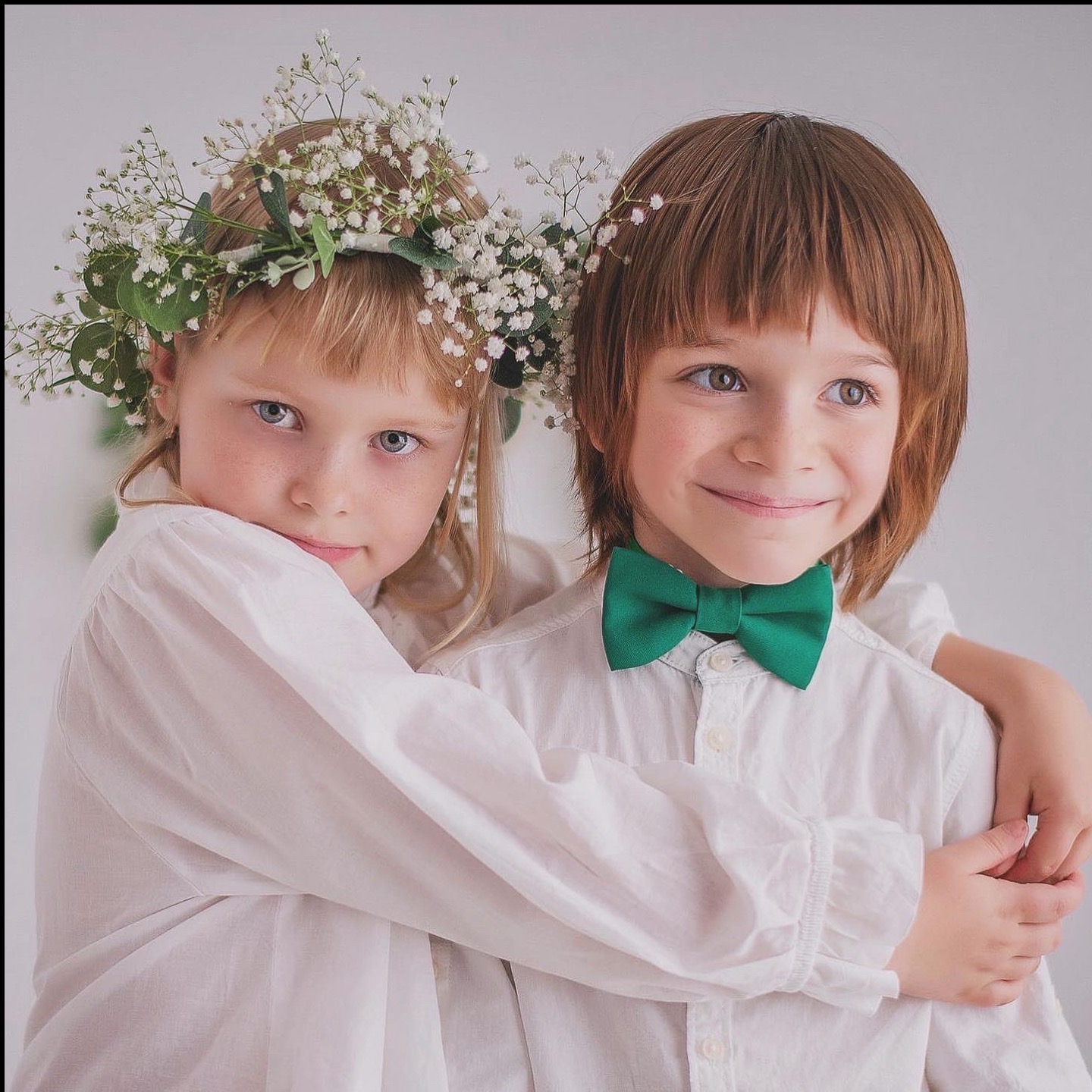 Portret rodzeństwa: dziewczynka w wianku z gipsówki obejmuje chłopca w białej koszuli i zielonej muszce na neutralnym tle. Delikatne światło, pastelowa kolorystyka.