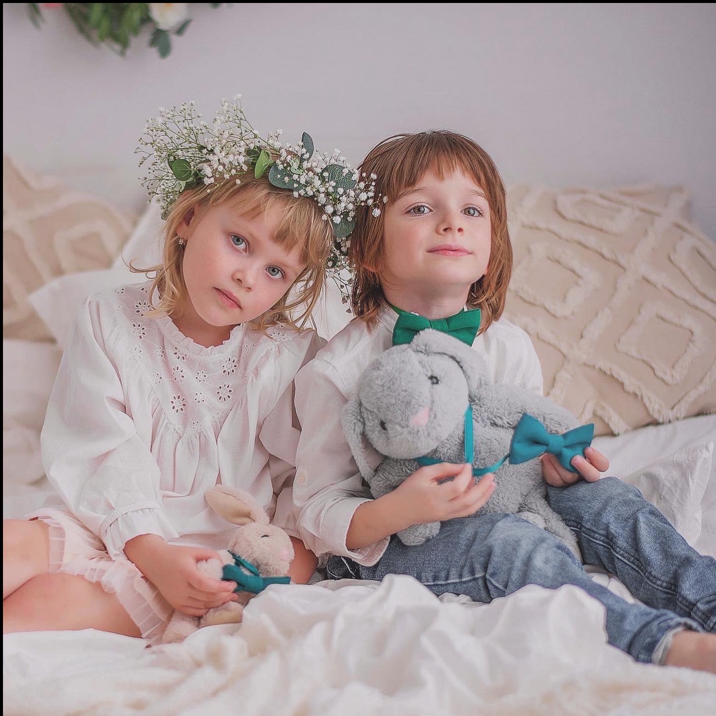 Dwoje dzieci siedzi na łóżku: dziewczynka w wianku z gipsówki i chłopiec w zielonej muszce, oboje trzymają pluszowe króliczki z zielonymi kokardkami.