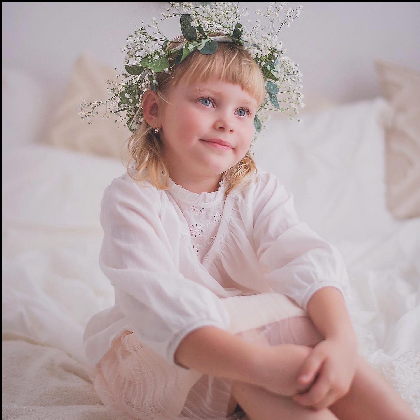 Urocza blond dziewczynka z wiankiem z gipsówki na głowie, ubrana w białą sukienkę, siedzi na białym łóżku, spoglądając w górę z lekkim uśmiechem. Subtelne oświetlenie.