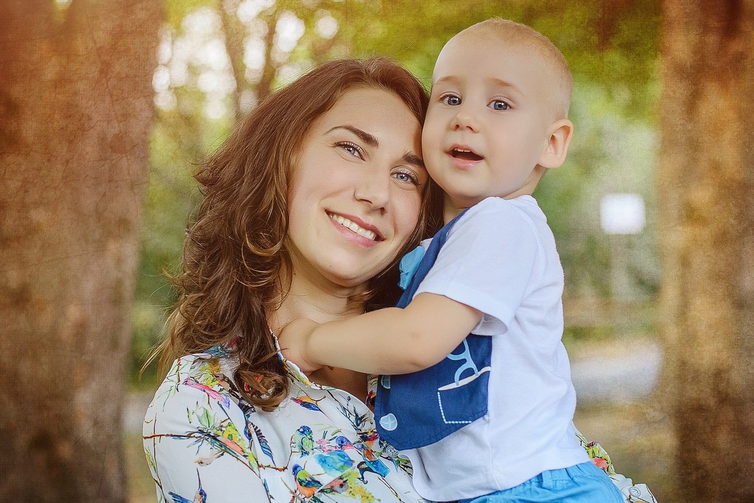 Portret uśmiechniętej matki z synkiem na rękach, w naturalnym oświetleniu, na tle drzew. Kobieta w bluzce w kolorowe ptaki, dziecko w białej koszulce i niebieskich spodenkach.