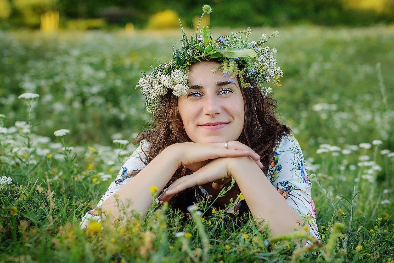Uśmiechnięta brunetka z wiankiem na głowie leży na łące pełnej białych i żółtych kwiatów. Delikatny makijaż, wzrok skierowany w obiektyw.