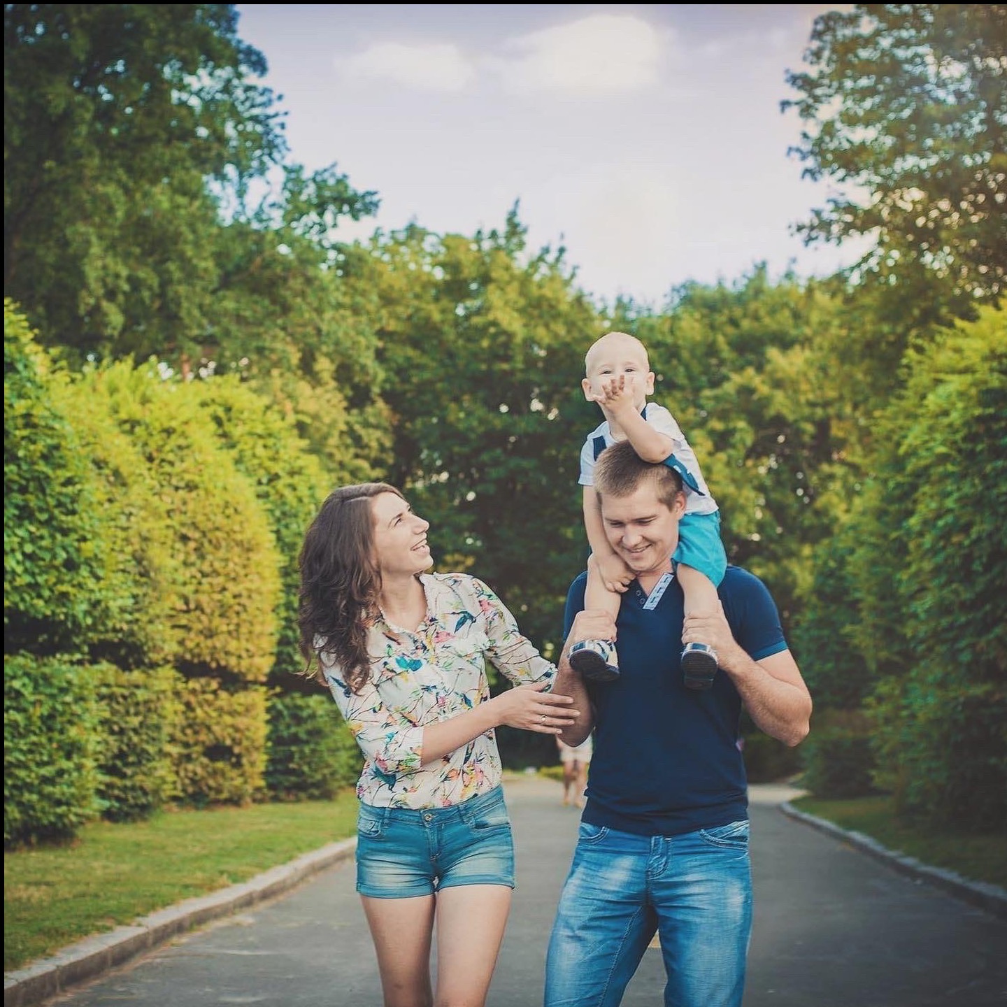 Uśmiechnięta rodzina: mama, tata z synkiem na barana, spacerują parkową alejką w słoneczny dzień. Dziecko radośnie macha ręką do obiektywu.
