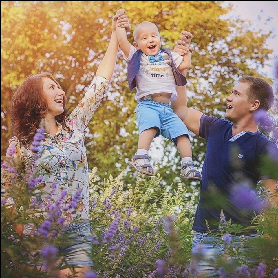 Radosna rodzina w letniej scenerii: mama trzyma za ręce podnoszone w górę dziecko, tata podtrzymuje je, wszyscy uśmiechnięci, otoczeni fioletowymi kwiatami.