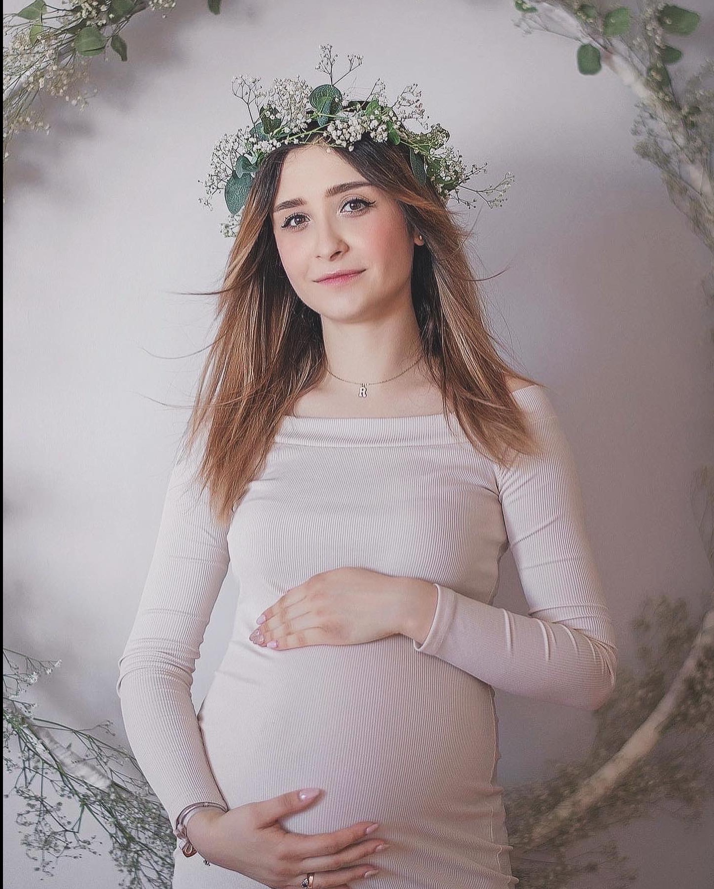 Portret uśmiechniętej kobiety w ciąży z wiankiem na głowie, w jasnej sukience, z rękami na brzuchu, na tle dekoracji z białych kwiatów i zielonych gałązek.