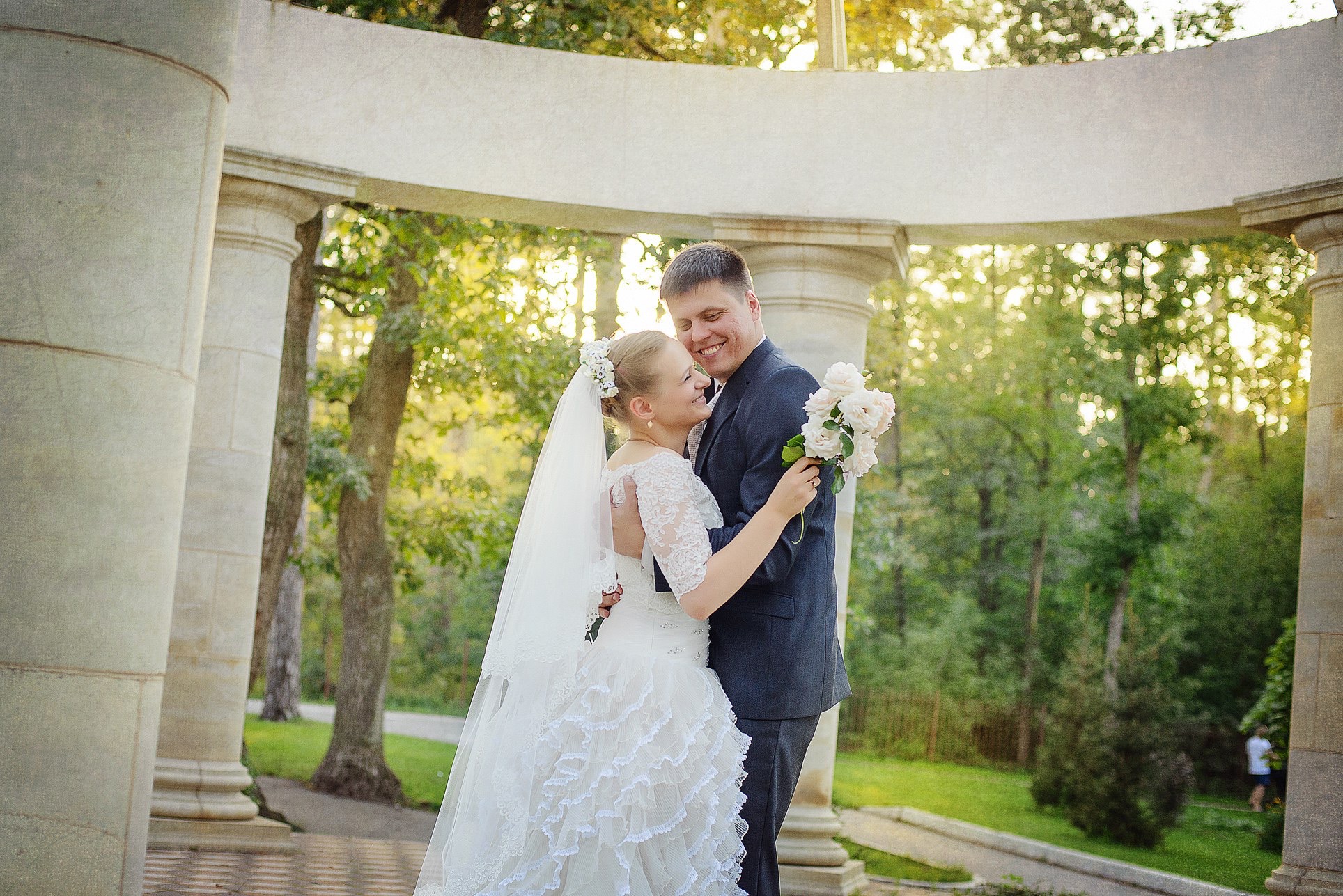 Uśmiechnięta para młoda w objęciach, ona w białej sukni z welonem, on w ciemnym garniturze, trzymają bukiet. W tle kolumnada i zieleń parku w ciepłym świetle.