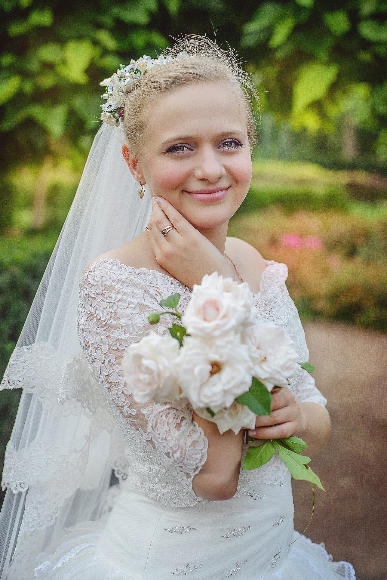 Uśmiechnięta panna młoda w koronkowej sukni, z bukietem róż w Lublinie, delikatnie dotyka twarzy. Romantyczna fotografia ślubna w plenerze.