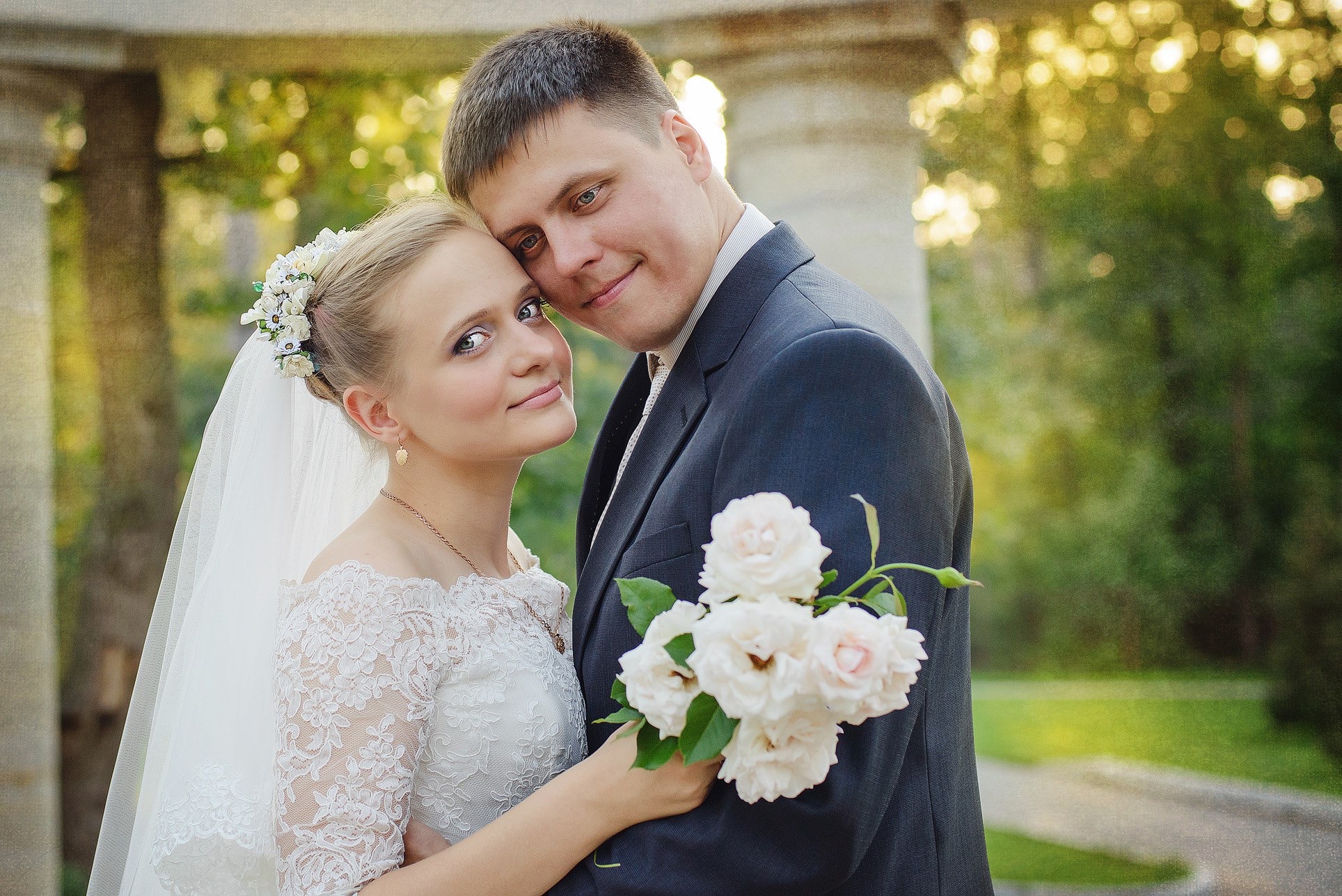 Uśmiechnięta para młoda w objęciach, panna młoda w koronkowej sukni i welonie, pan młody w ciemnym garniturze trzyma bukiet białych róż na tle zieleni.