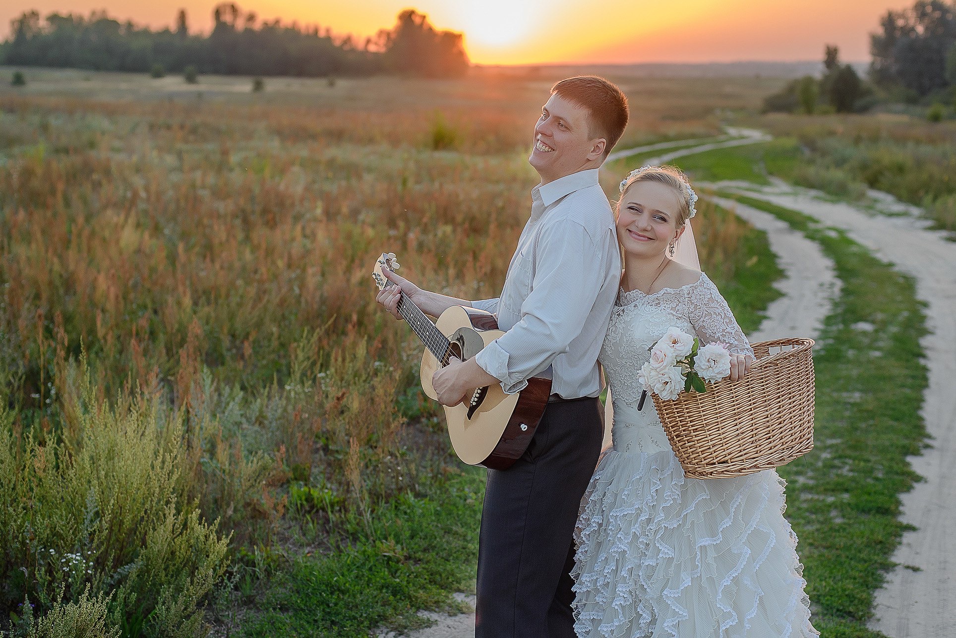 Para młoda w plenerze: on gra na gitarze, ona trzyma koszyk z kwiatami. Romantyczna sesja ślubna na tle zachodzącego słońca i polnej drogi.