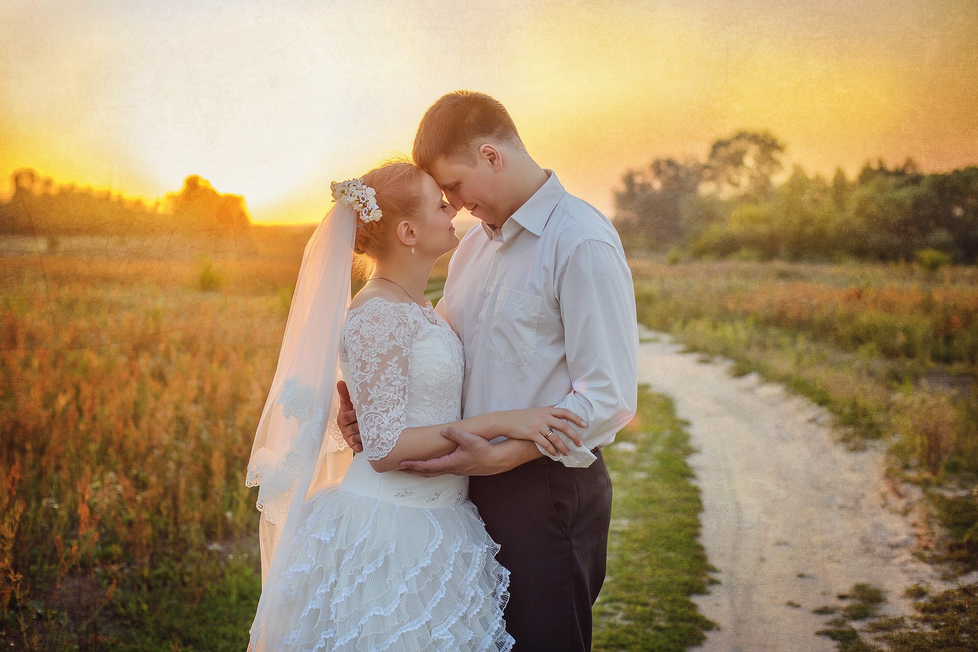 Para młoda w objęciach na polnej drodze o zachodzie słońca. Panna młoda w koronkowej sukni i welonie z kwiatami, pan młody w jasnej koszuli. Romantyczna atmosfera.