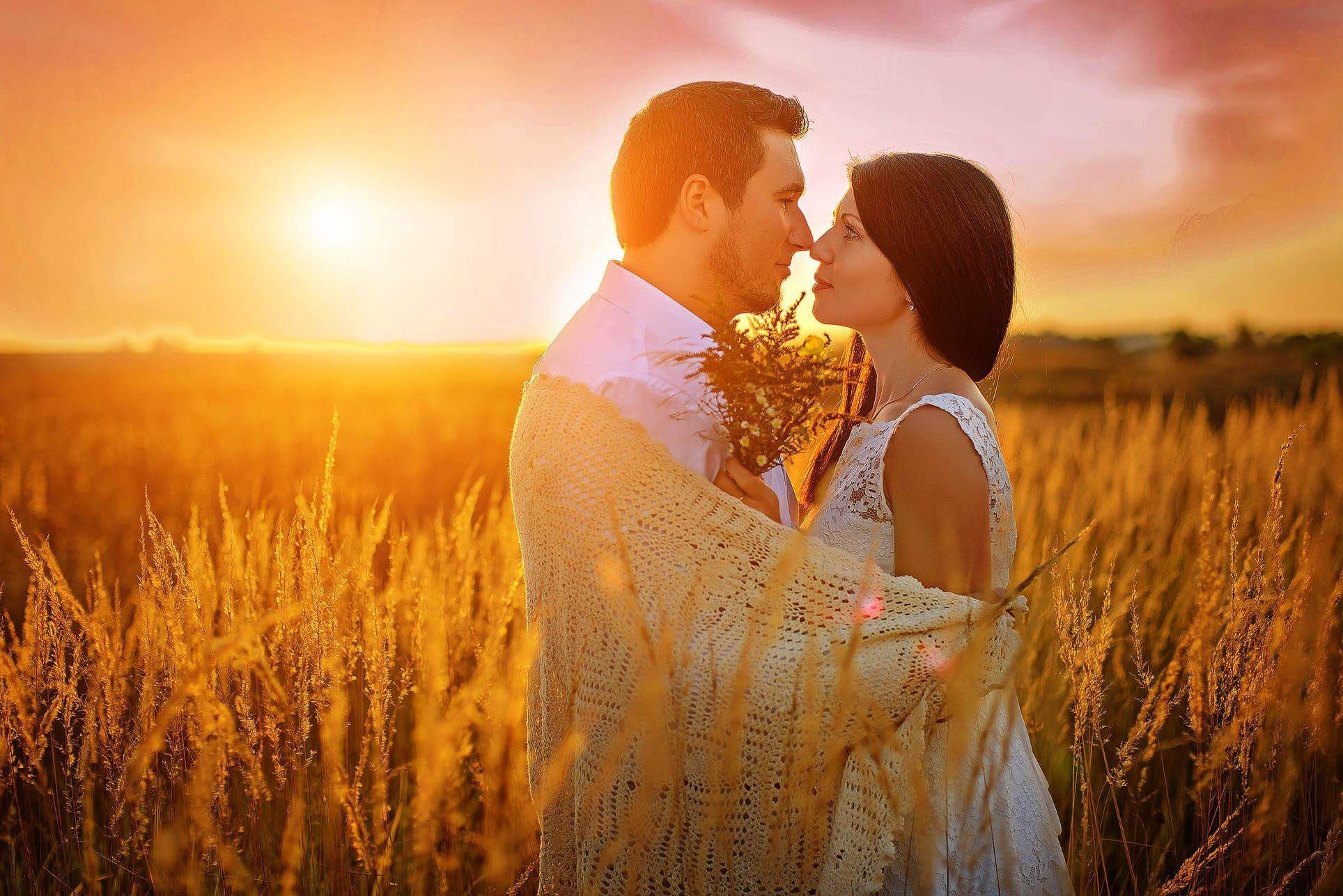 Zakochana para w objęciach, na tle zachodzącego słońca i pola złocistych traw. Mężczyzna z bukietem, oboje otuleni kremowym pledem.