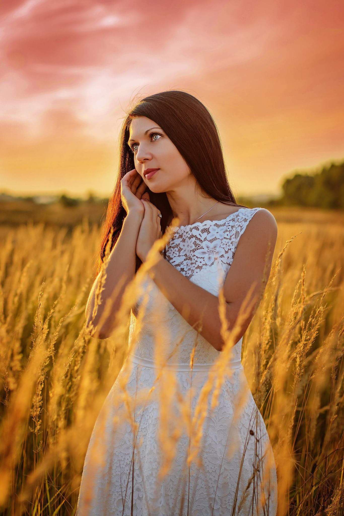 Kobieta w białej koronkowej sukience stoi na polu trawy w ciepłym, zachodzącym słońcu, z rękami delikatnie dotykającymi twarzy i zamyślonym spojrzeniem w bok.