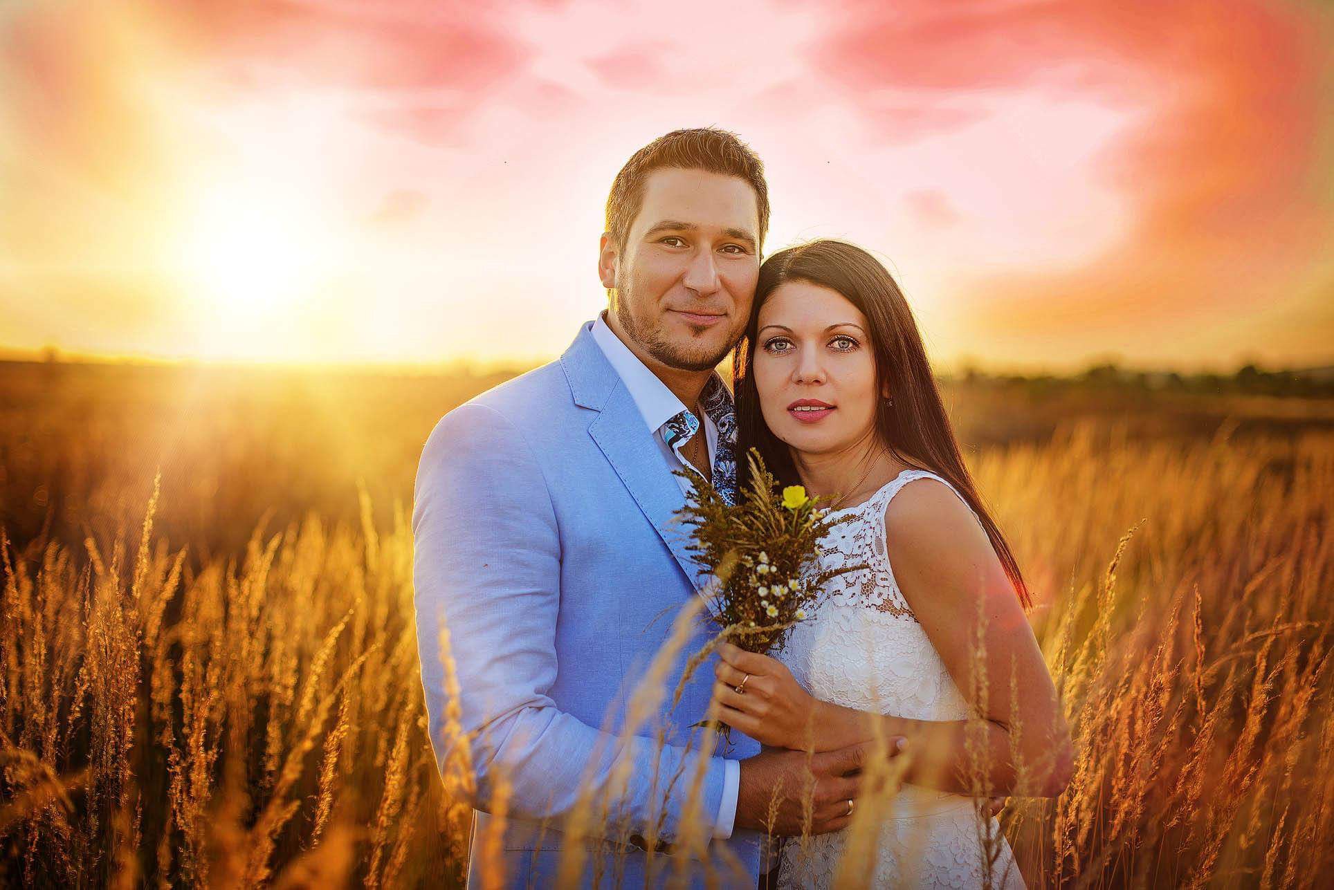 Para w objęciach na tle zachodzącego słońca w polu. Kobieta w koronkowej sukience trzyma bukiecik polnych kwiatów, mężczyzna w błękitnej marynarce.