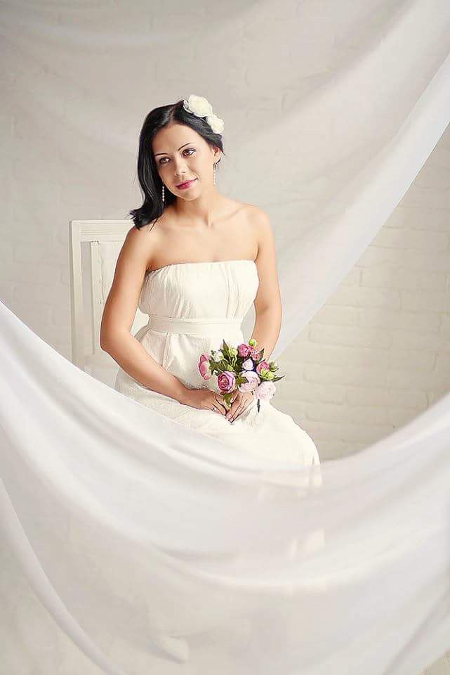 Uśmiechnięta panna młoda w białej sukni bez ramiączek, siedząca na krześle, trzyma bukiet różowych kwiatów. Włosy upięte kwiatami, delikatna biżuteria.