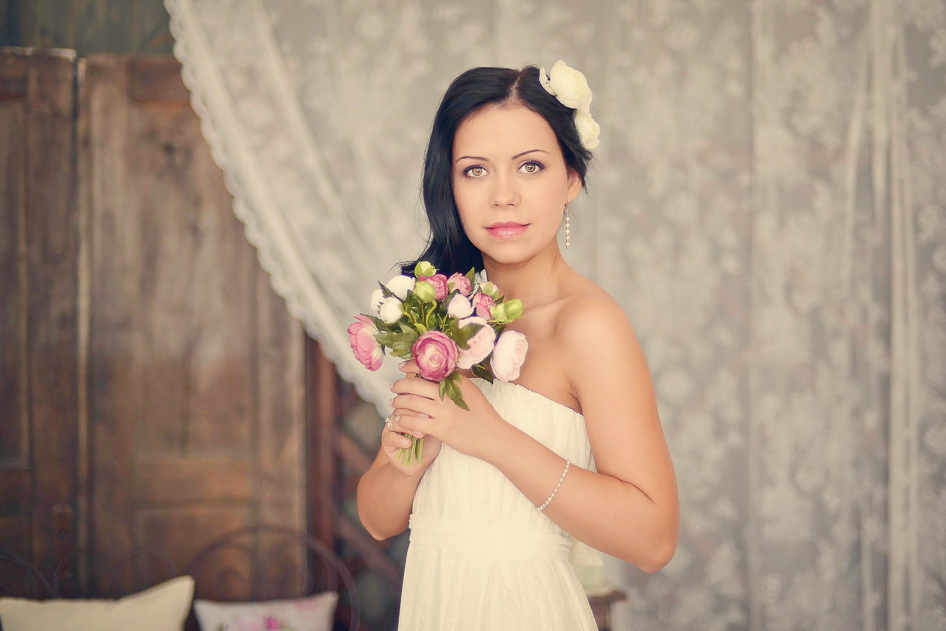 Portret kobiety w białej sukni, z kwiatami we włosach i bukietem w dłoniach, na tle vintage drzwi i zasłon. Delikatne oświetlenie, subtelny makijaż.