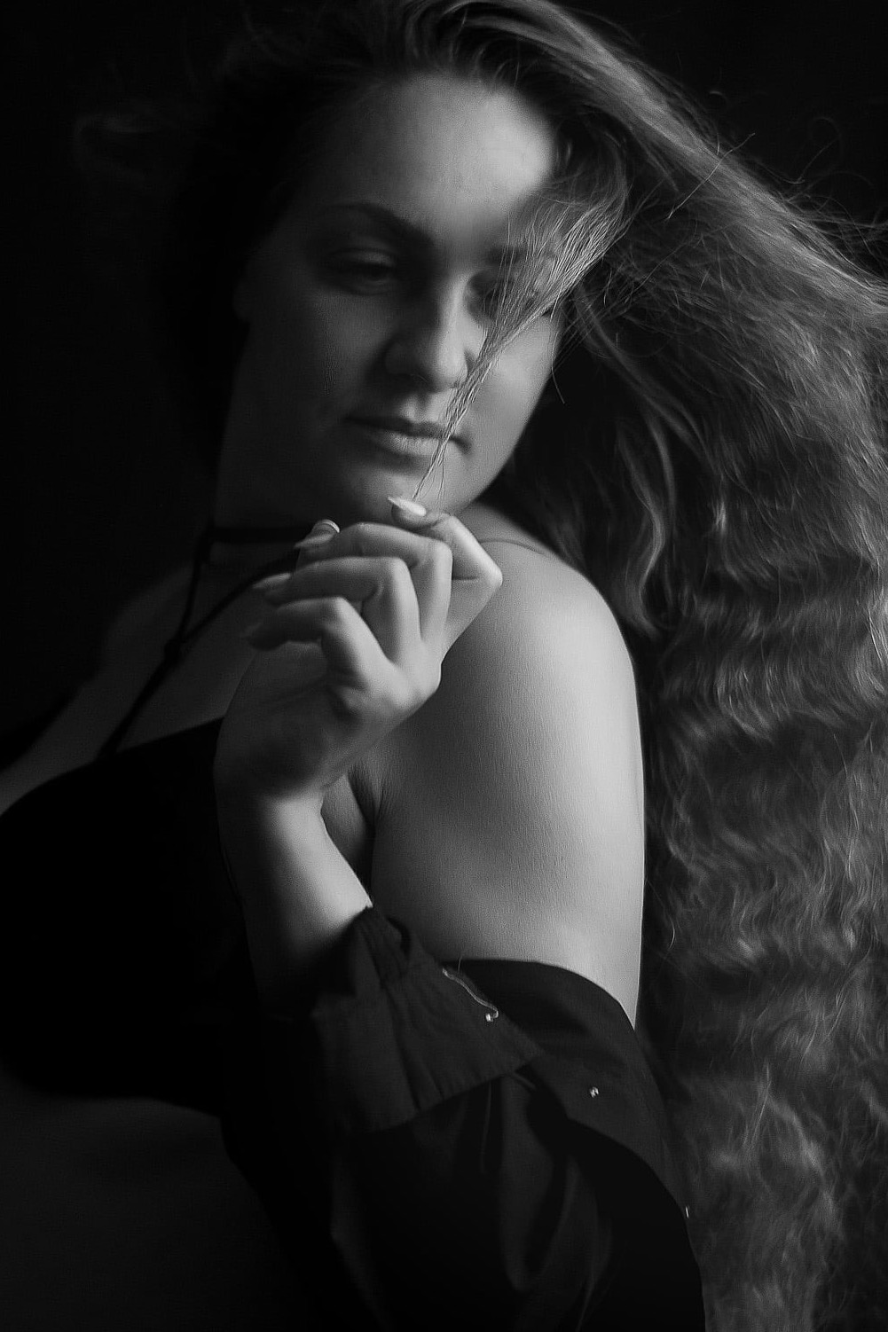 Czarno-biały portret kobiety z długimi, falującymi włosami, zamyślona poza, delikatne światło podkreśla rysy twarzy i fakturę włosów.