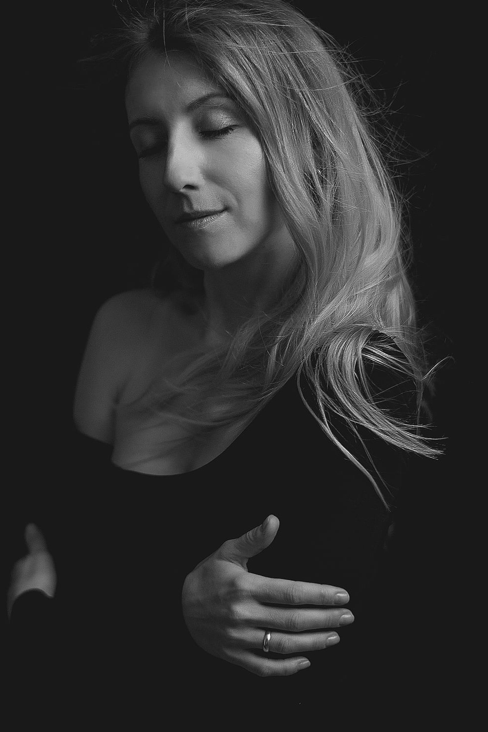 Czarno-biały portret kobiety z zamkniętymi oczami, delikatnym uśmiechem i dłonią z pierścionkiem na ciemnym tle. Włosy opadają na ramię, tworząc miękkie fale.