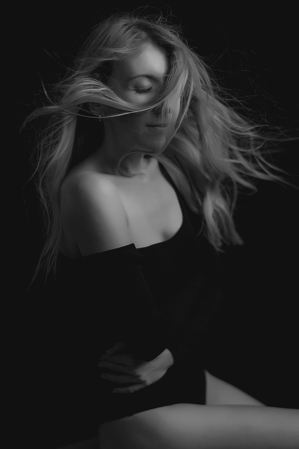 Czarno-biały portret kobiety z rozwianymi włosami, zamkniętymi oczami i delikatnym uśmiechem, na ciemnym tle. Subtelne światło podkreśla rysy twarzy i fakturę włosów.