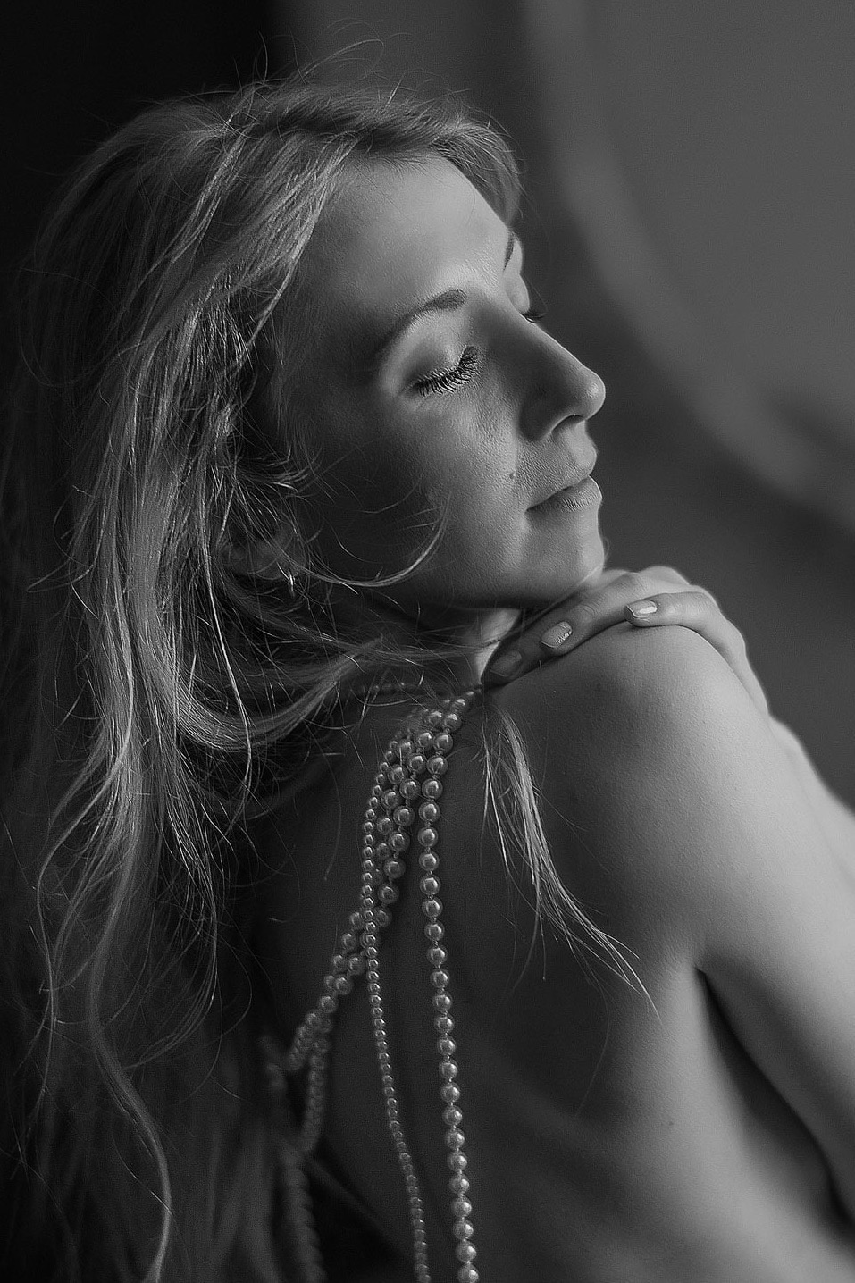 Czarno-biały portret profilowy kobiety z perłami na ramieniu, delikatne światło, zamknięte oczy, subtelny makijaż, artystyczny klimat.