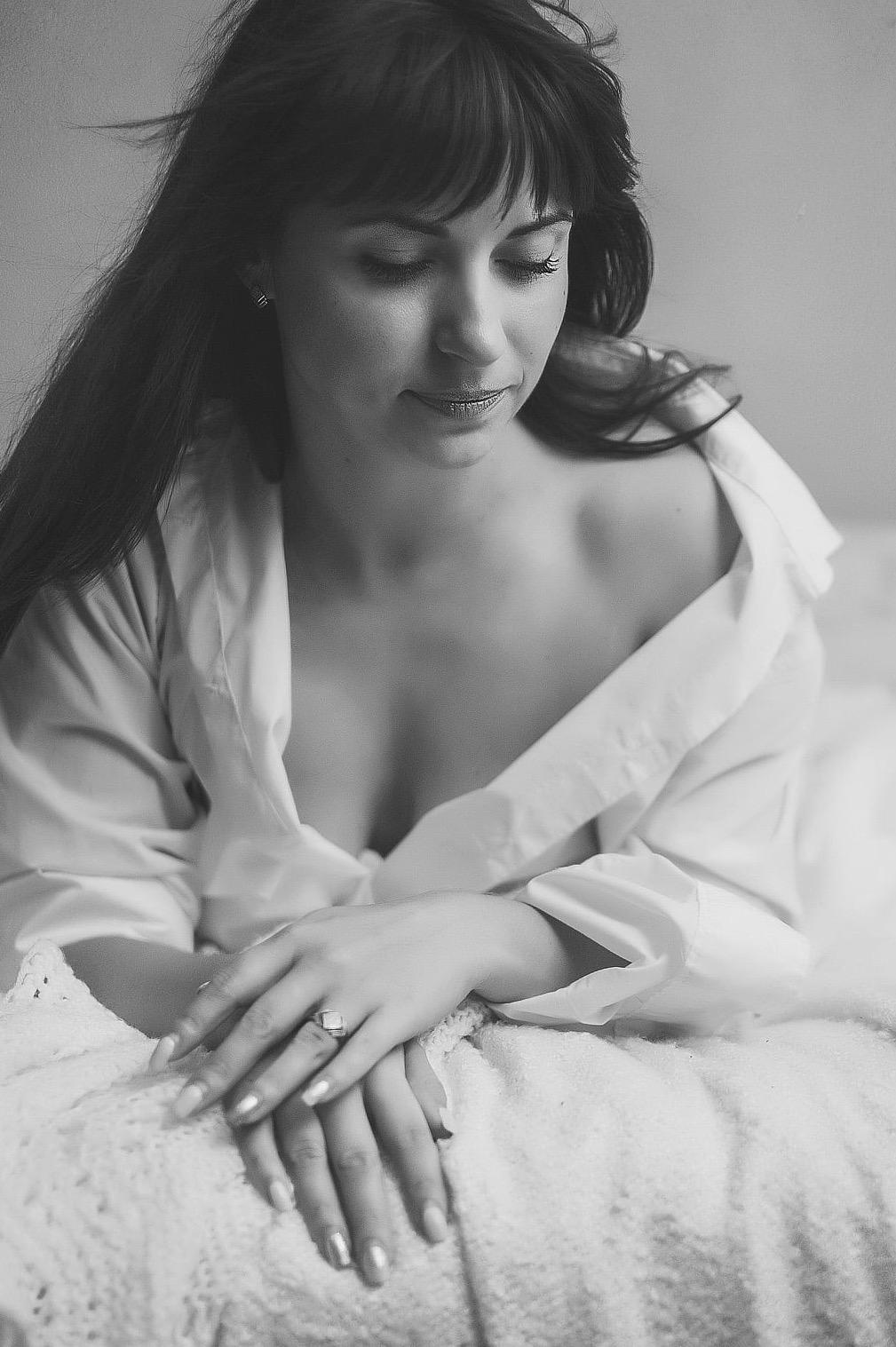 Portret kobiety w białej koszuli, z delikatnym uśmiechem i pierścionkiem na palcu, w czarno-białej tonacji, z miękkim światłem padającym na twarz.