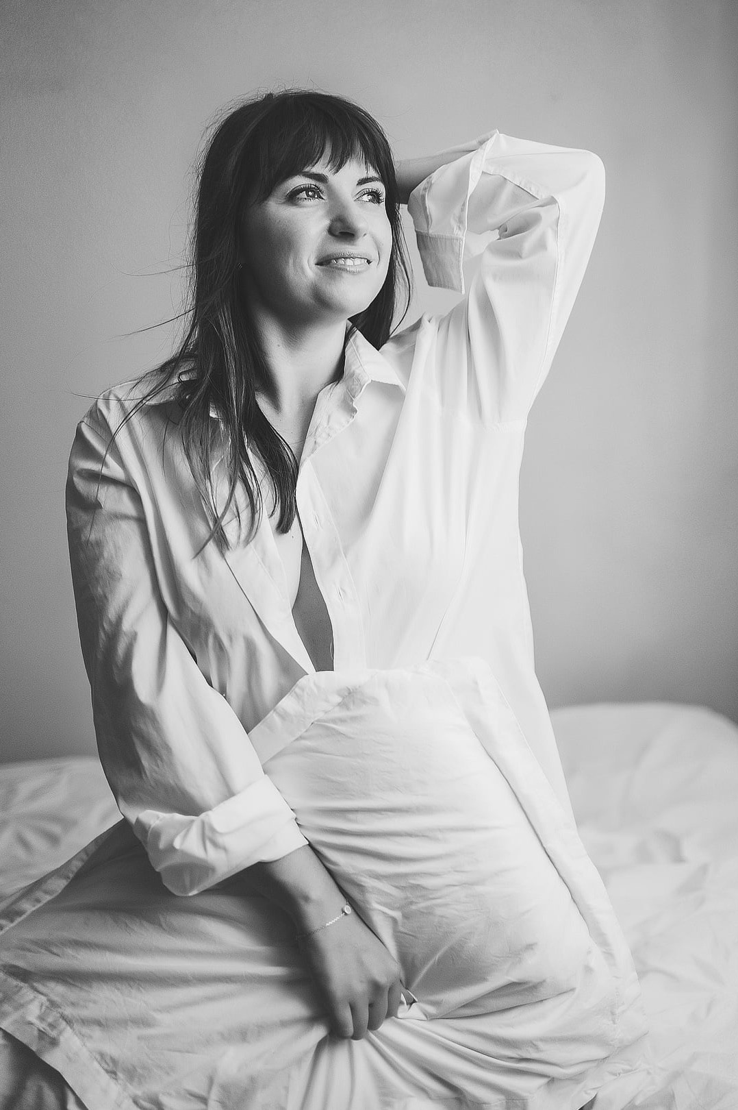 Portret kobiety w białej koszuli, siedzącej na łóżku i trzymającej poduszkę. Zdjęcie czarno-białe, z delikatnym uśmiechem i spojrzeniem w górę.