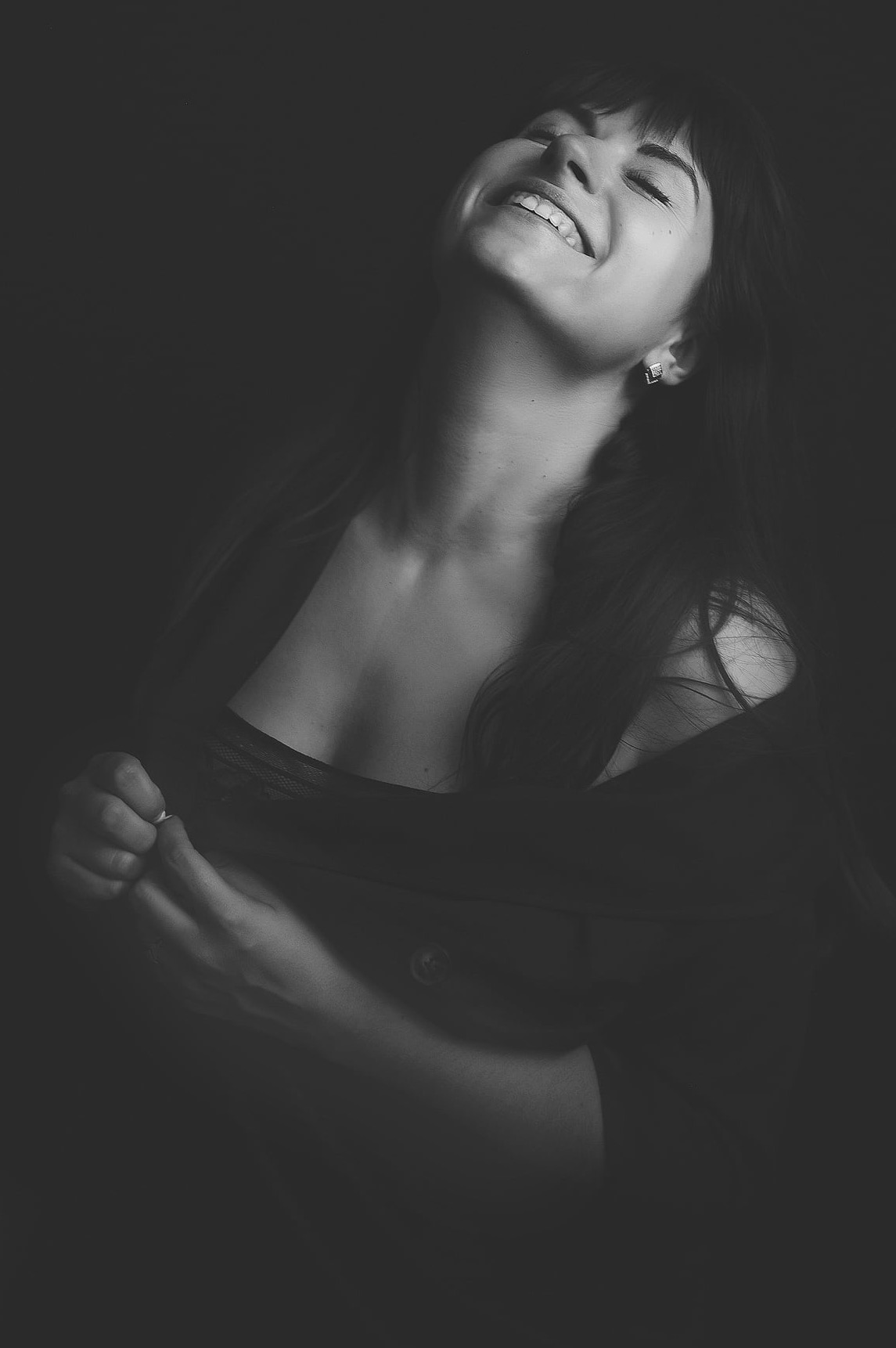 Czarno-biały portret uśmiechniętej kobiety z zamkniętymi oczami, głowa odchylona do tyłu, z widocznym dekoltem i delikatną biżuterią w uchu, na ciemnym tle.