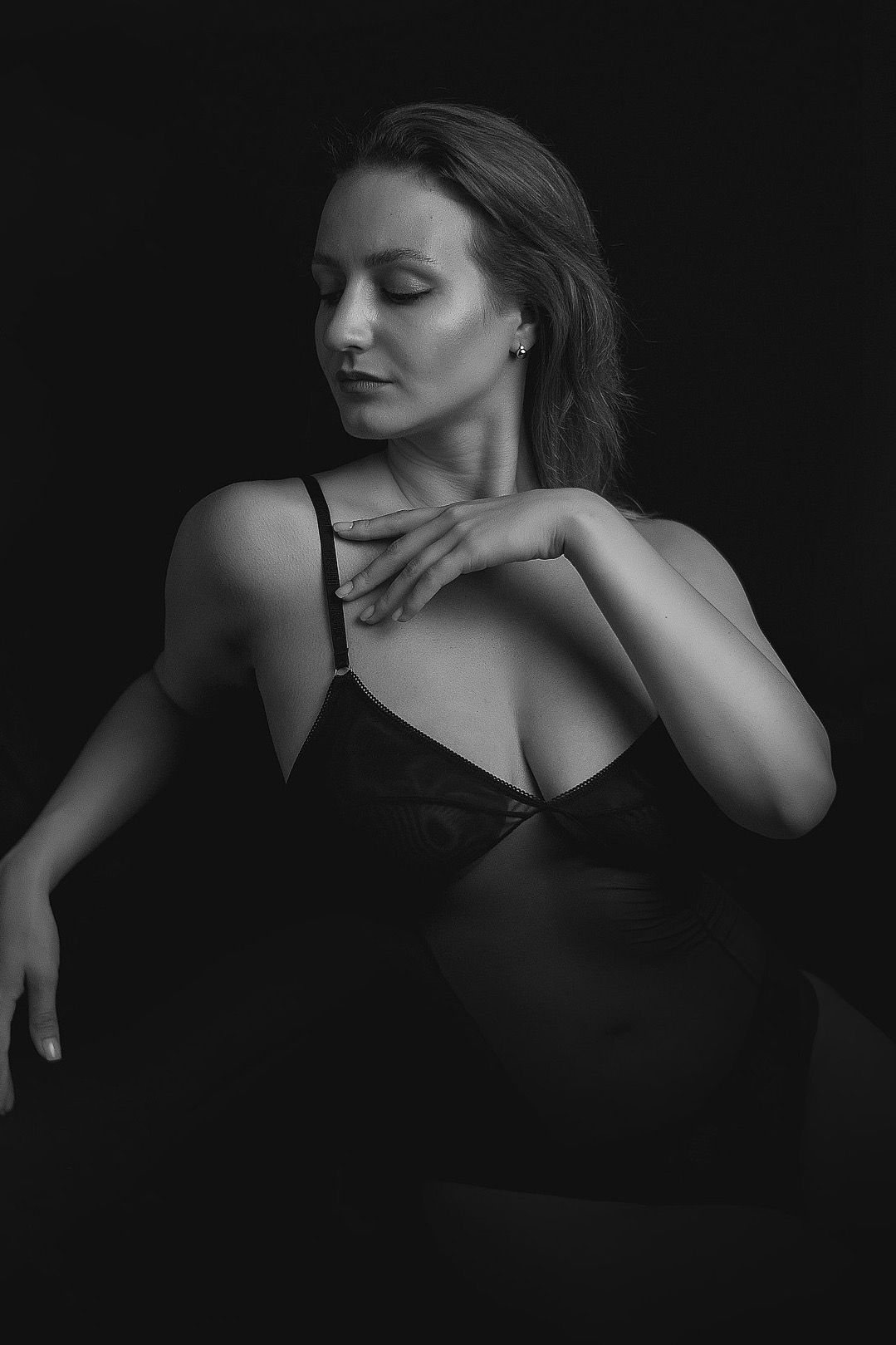 Czarno-białe, artystyczne ujęcie kobiety w bieliźnie, z delikatnym oświetleniem podkreślającym kontury ciała na ciemnym tle. Zamknięte oczy i subtelny gest dłoni dodają intymności.