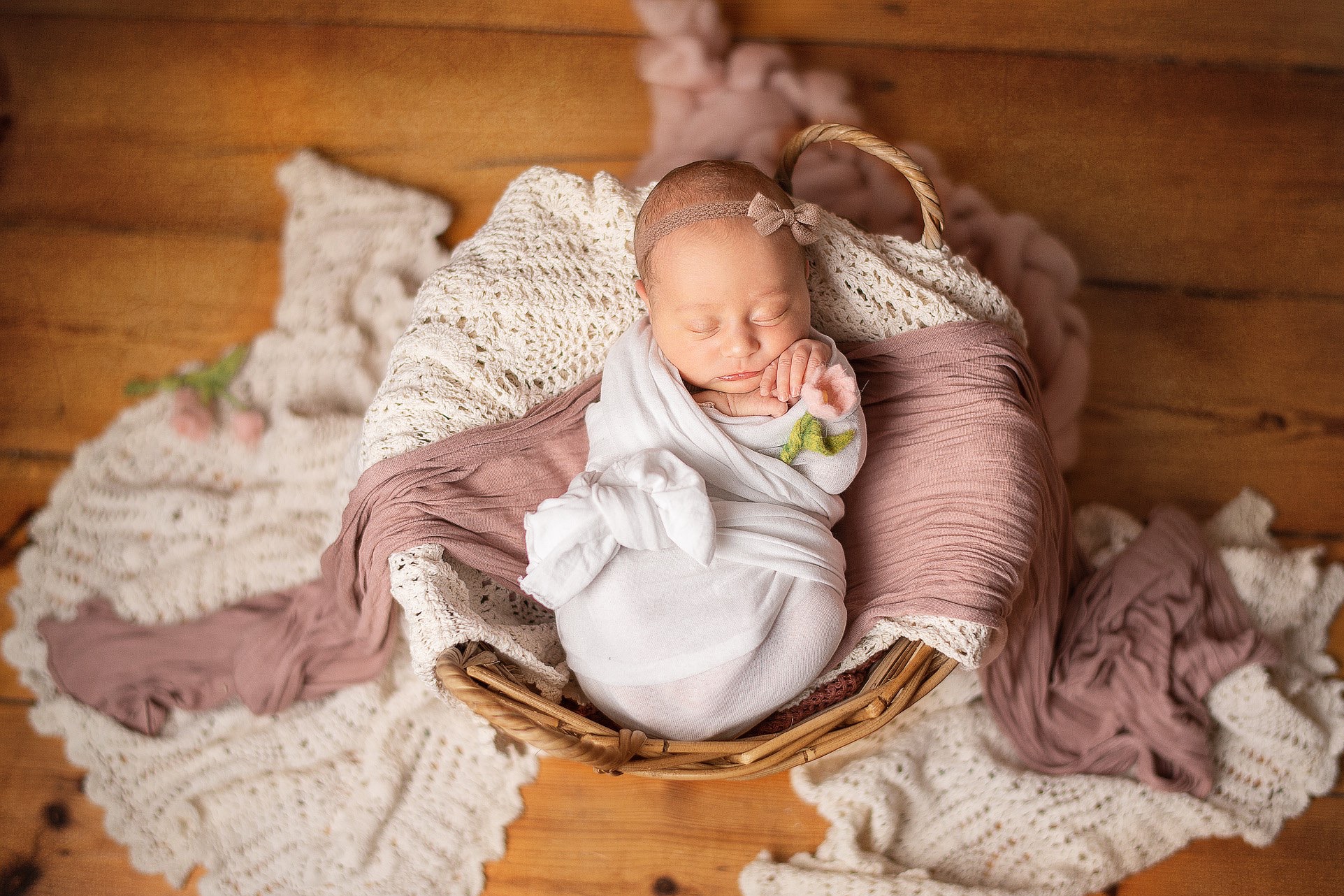 Noworodek zawinięty w biały materiał, śpiący w wiklinowym koszu na drewnianym tle, z różową chustą i koronkową serwetką. Dziecko ma opaskę z kokardką.