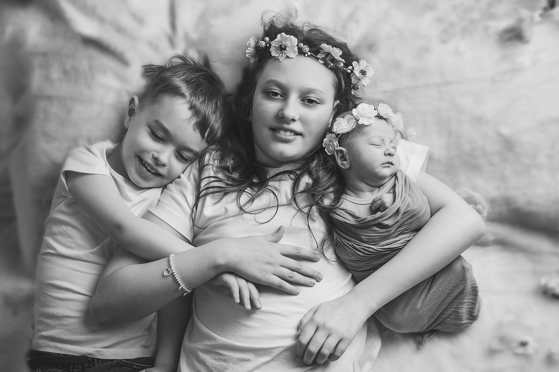 Czarno-białe zdjęcie z góry przedstawia trójkę dzieci: niemowlę owinięte w materiał i z wiankiem, nastolatkę z wiankiem i chłopca obejmującego nastolatkę.