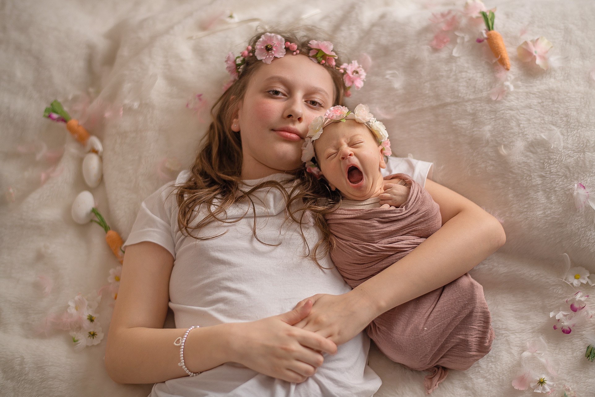 Uśmiechnięta nastolatka z wiankiem obejmuje noworodka w chuście, który ziewa. W tle białe futro, kwiaty i dekoracyjne marchewki tworzą wiosenną scenerię.